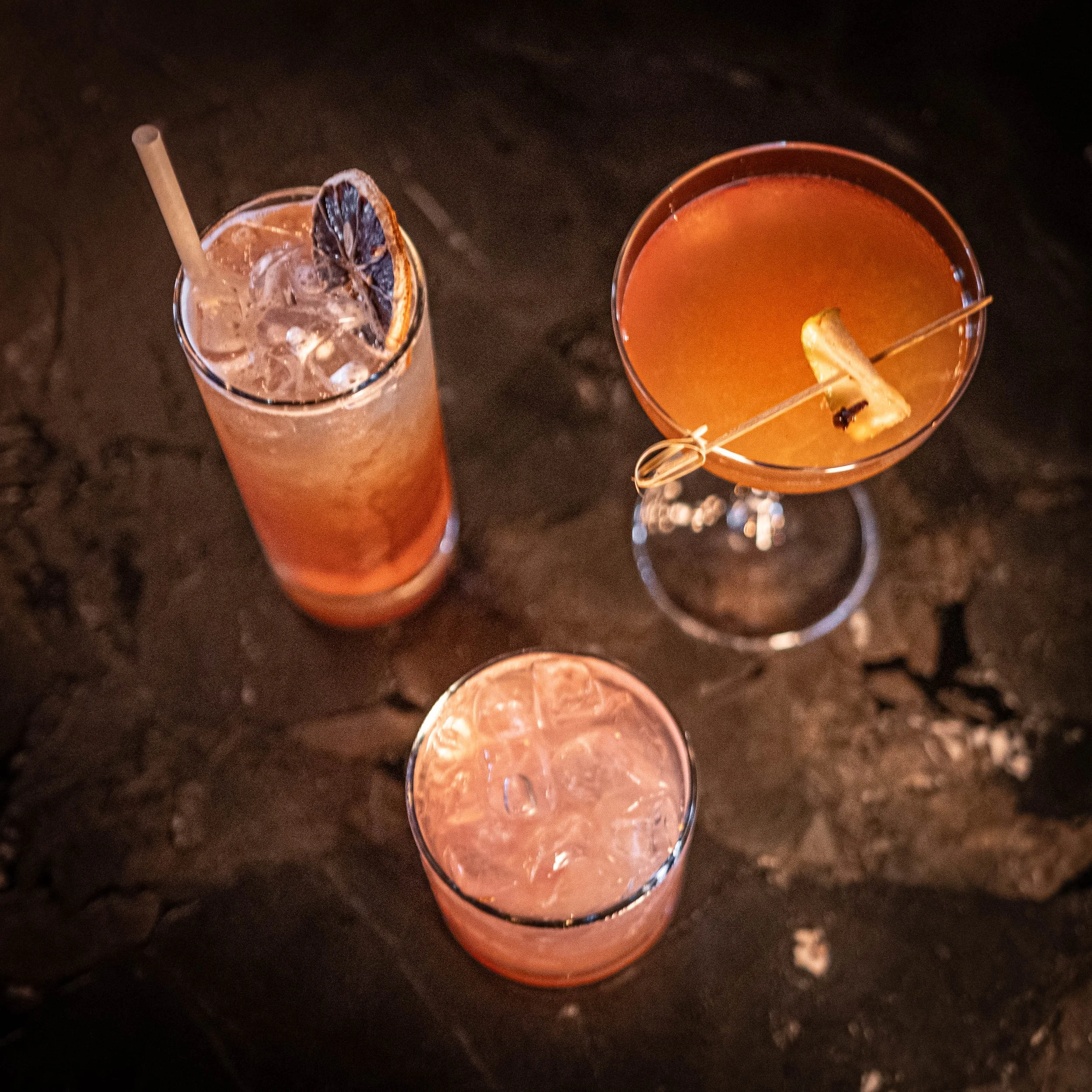 Three cocktails on a dark table, one tall glass with ice and a lemon slice, another in a martini glass with a lemon twist, and a third filled with a pinkish drink and ice.