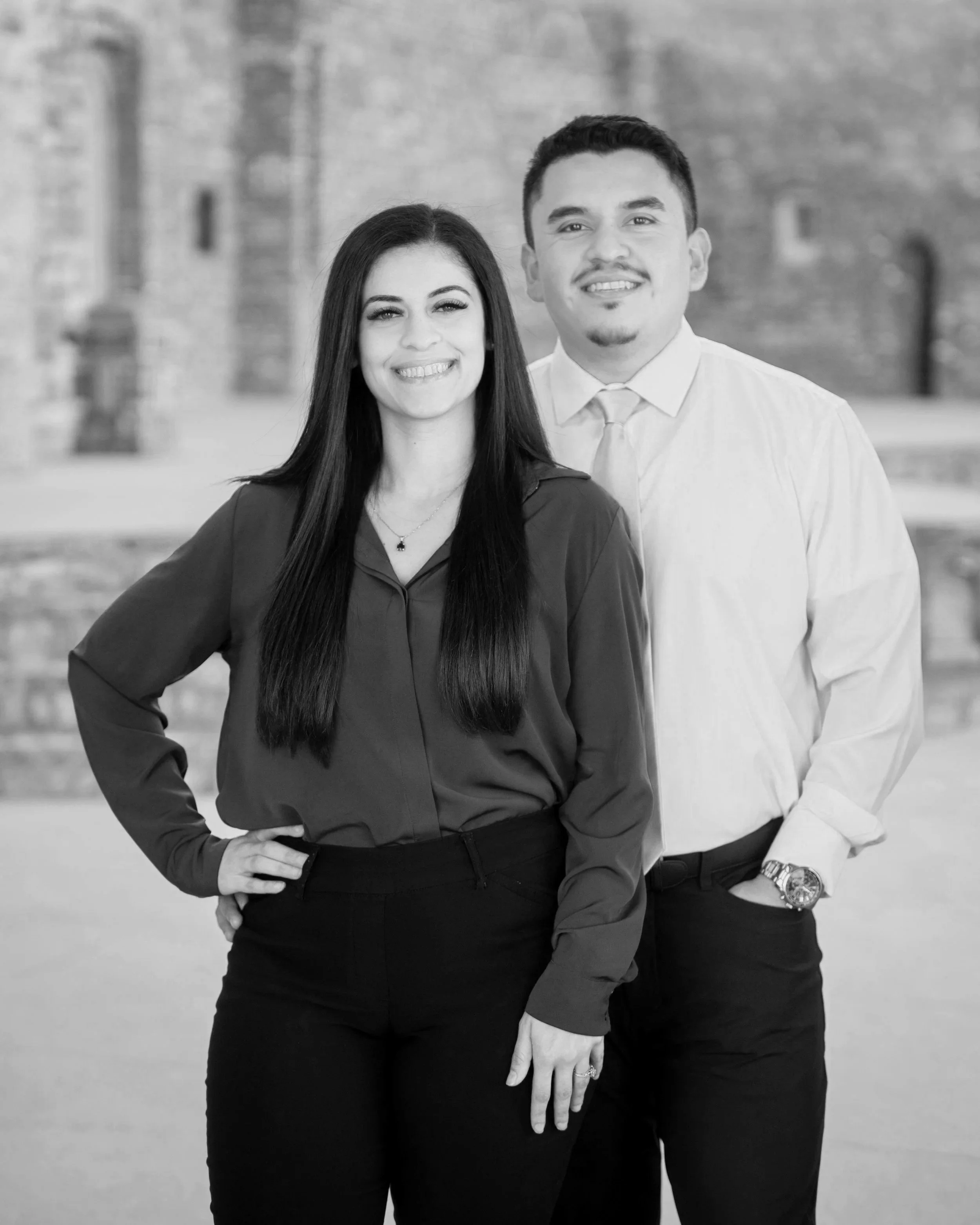 A black and white photo of a smiling woman and man standing outdoors with a brick building in the background. The woman has long dark hair, wears a button-down shirt, and has her hand on her hip. The man has short dark hair, wears a dress shirt with rolled-up sleeves, and has his hands in his pockets.