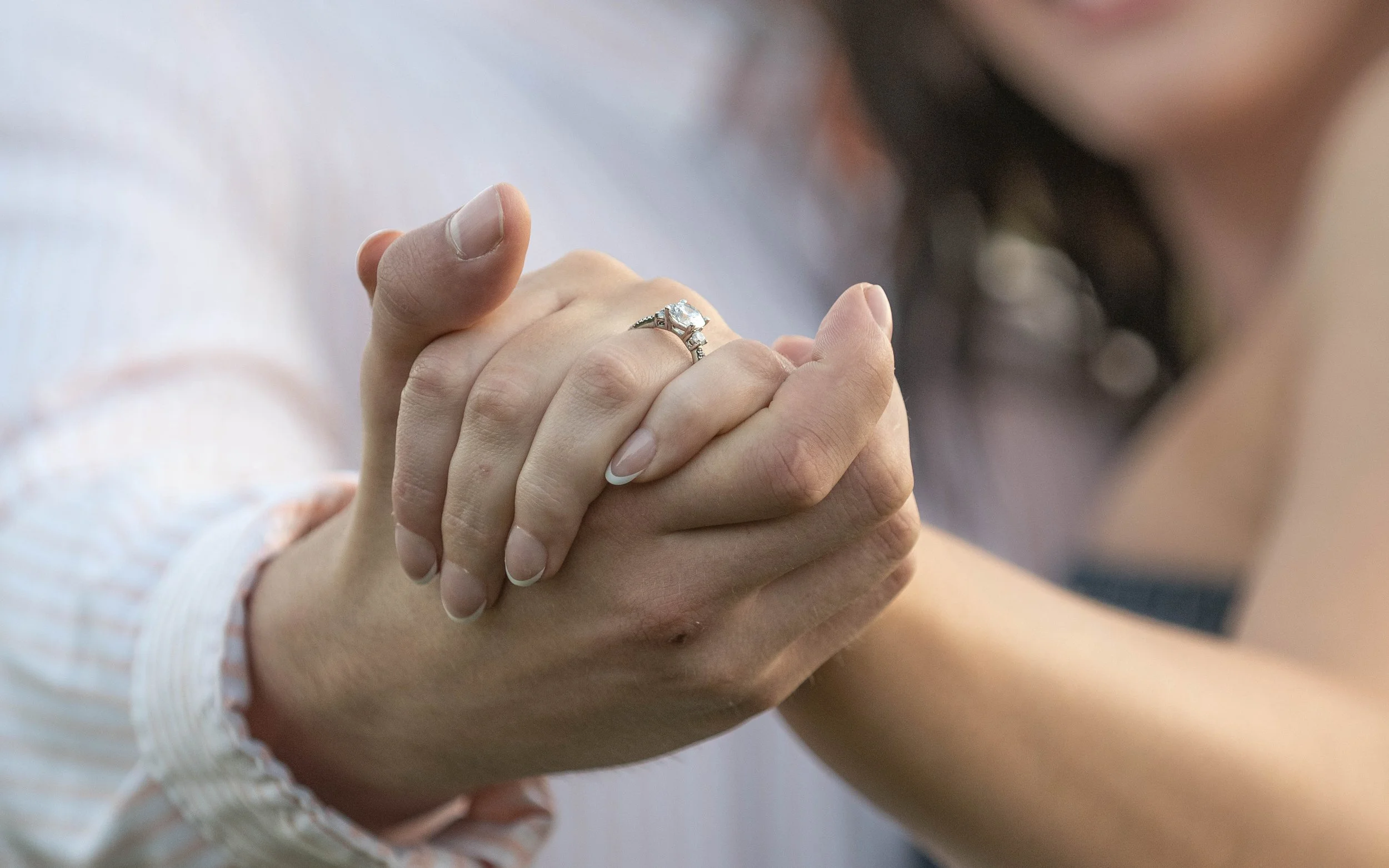 Close-up of a woman and a man holding hands, with the woman showing her engagement ring.