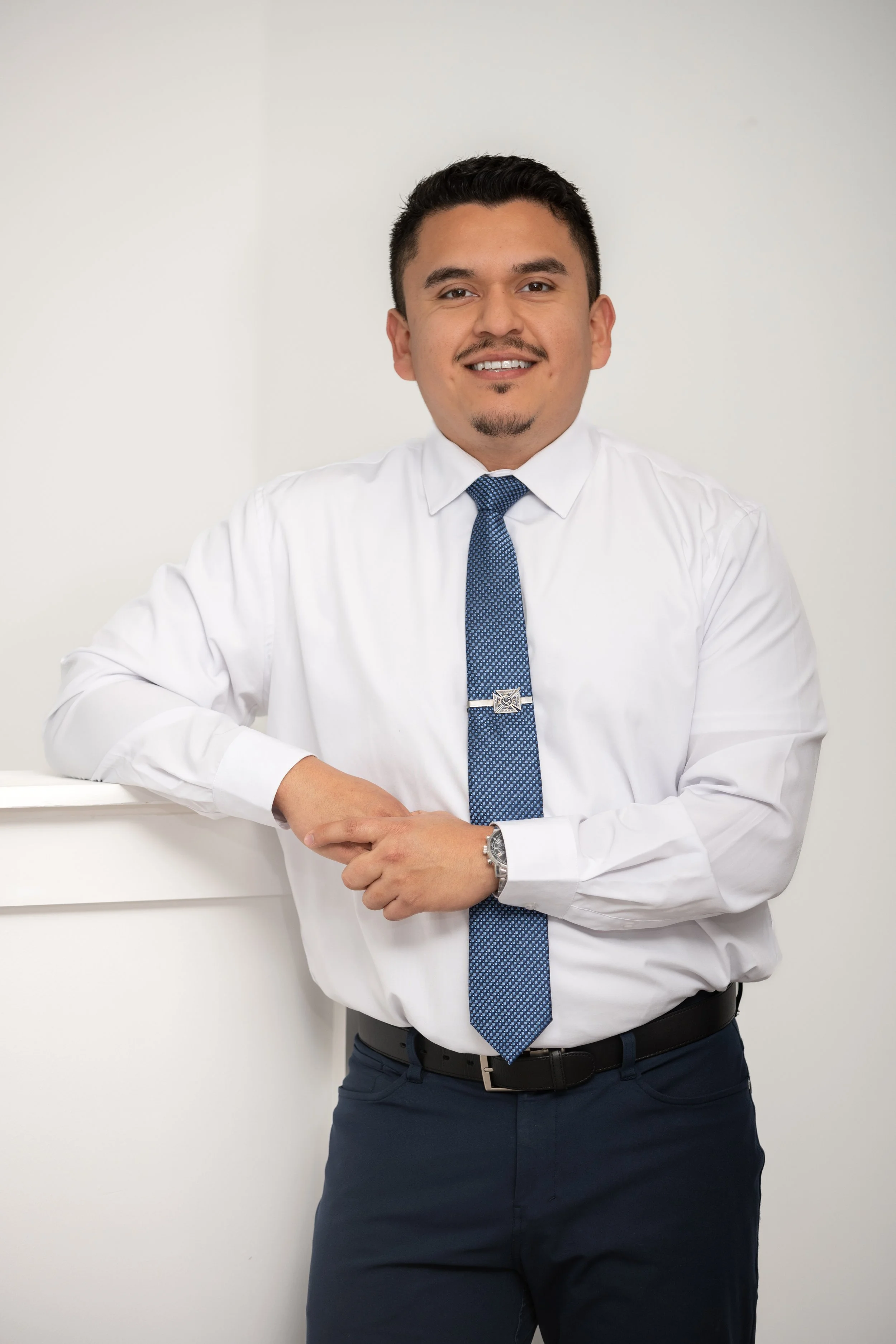 A man dressed in a white shirt, blue tie, and dark pants leaning against a white surface, smiling at the camera with a neutral background.