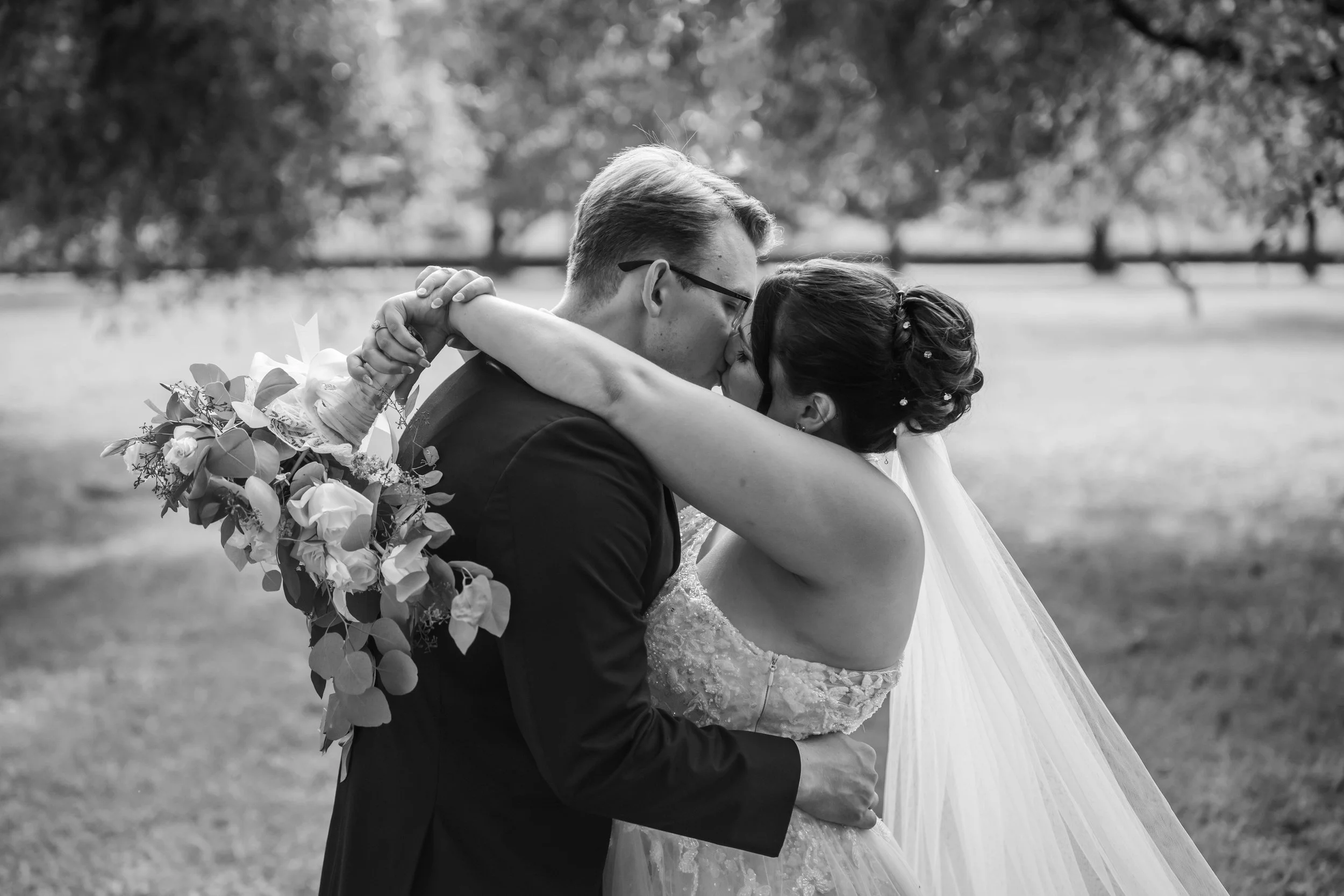 A black-and-white photo of a bride and groom kissing outdoors, with the bride holding a bouquet of flowers behind the groom's back.