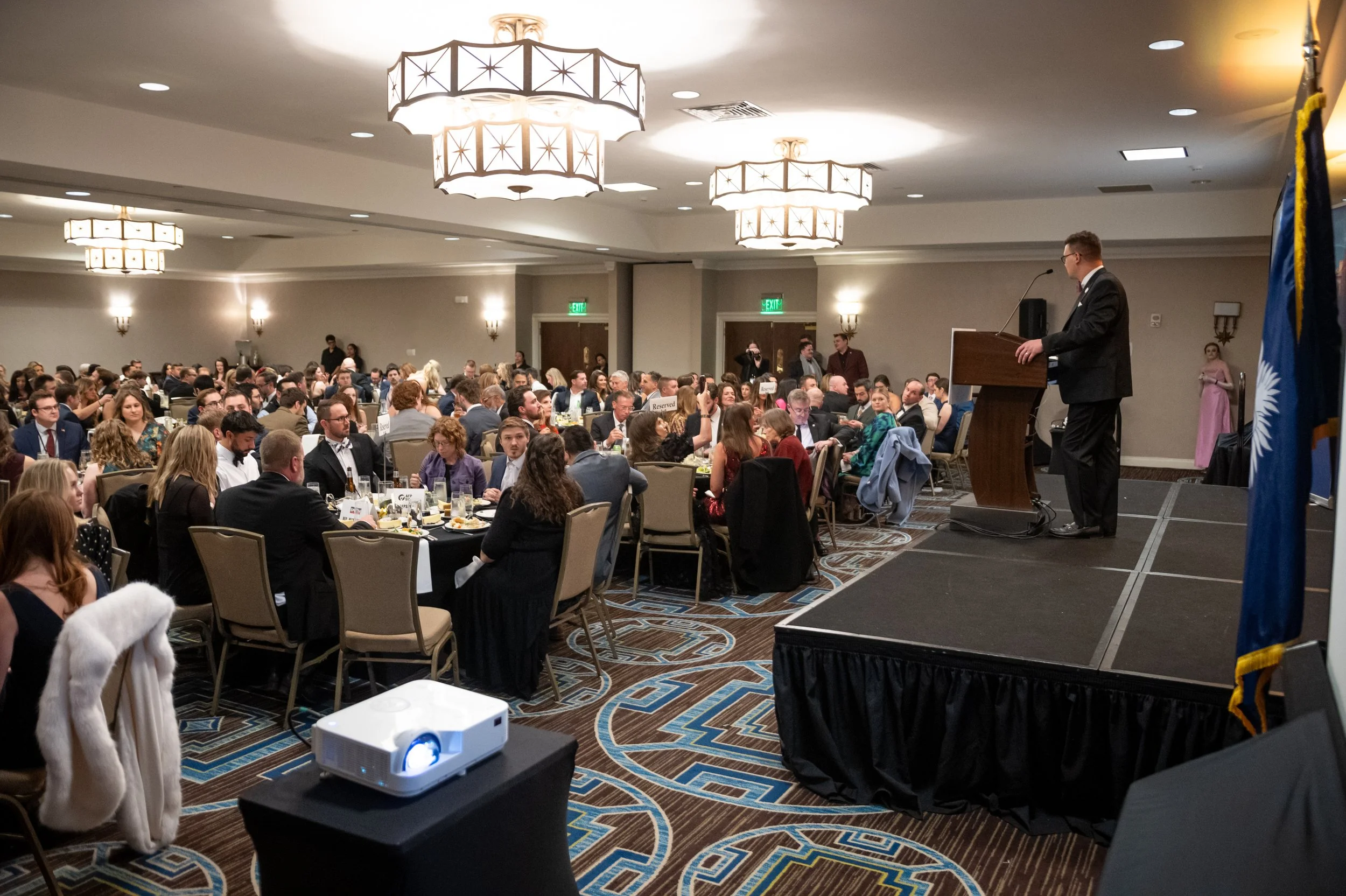A man in a suit giving a speech at a podium during a formal event in a large banquet hall filled with seated guests dining and listening.
