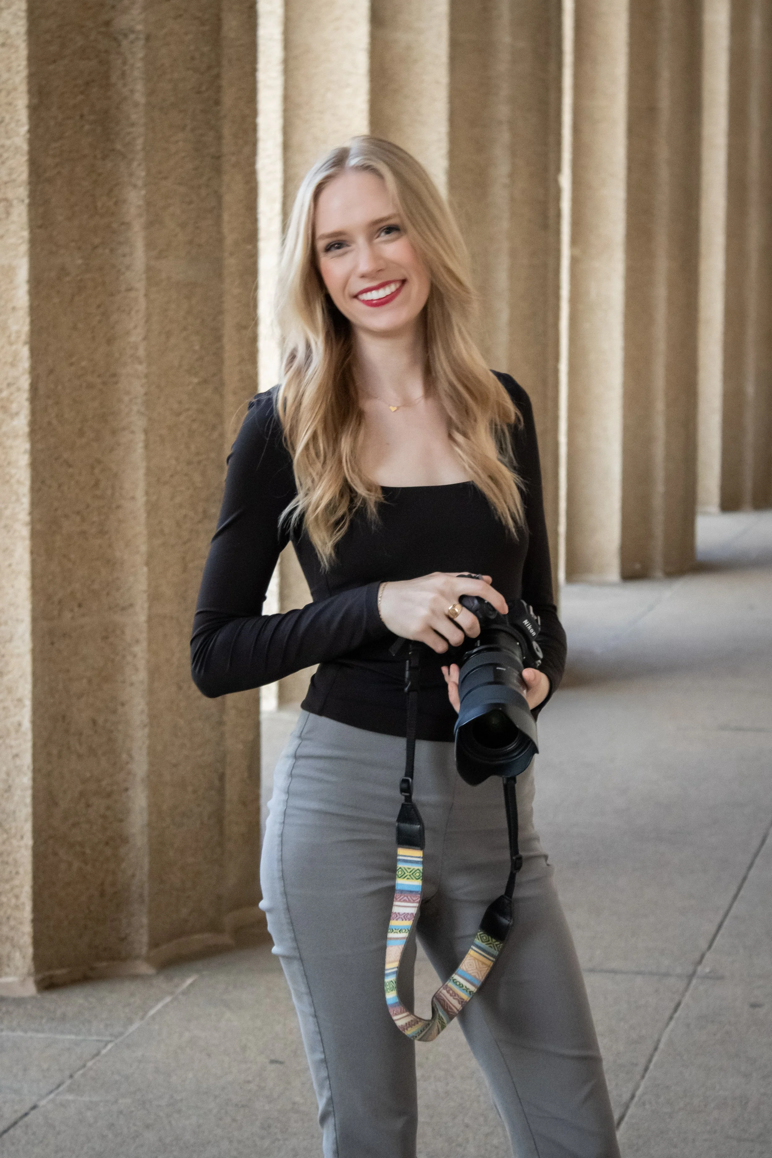 A young woman with blonde hair, smiling, holding a camera, standing outside near beige stone columns.