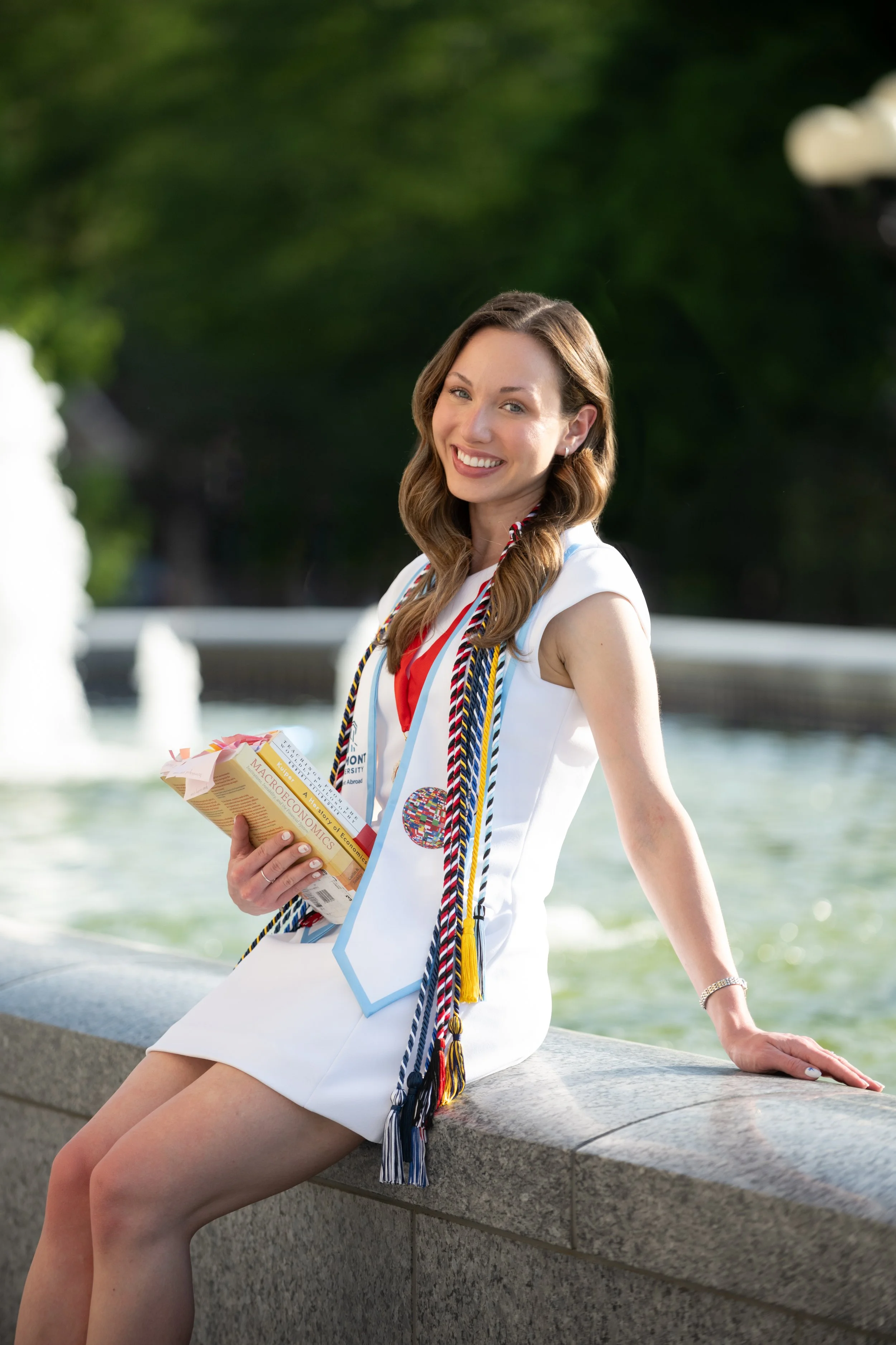 A young woman in a white sleeveless dress is sitting on a stone ledge near a fountain, wearing graduation cords and holding a book and papers, smiling outdoors with greenery and water in the background.