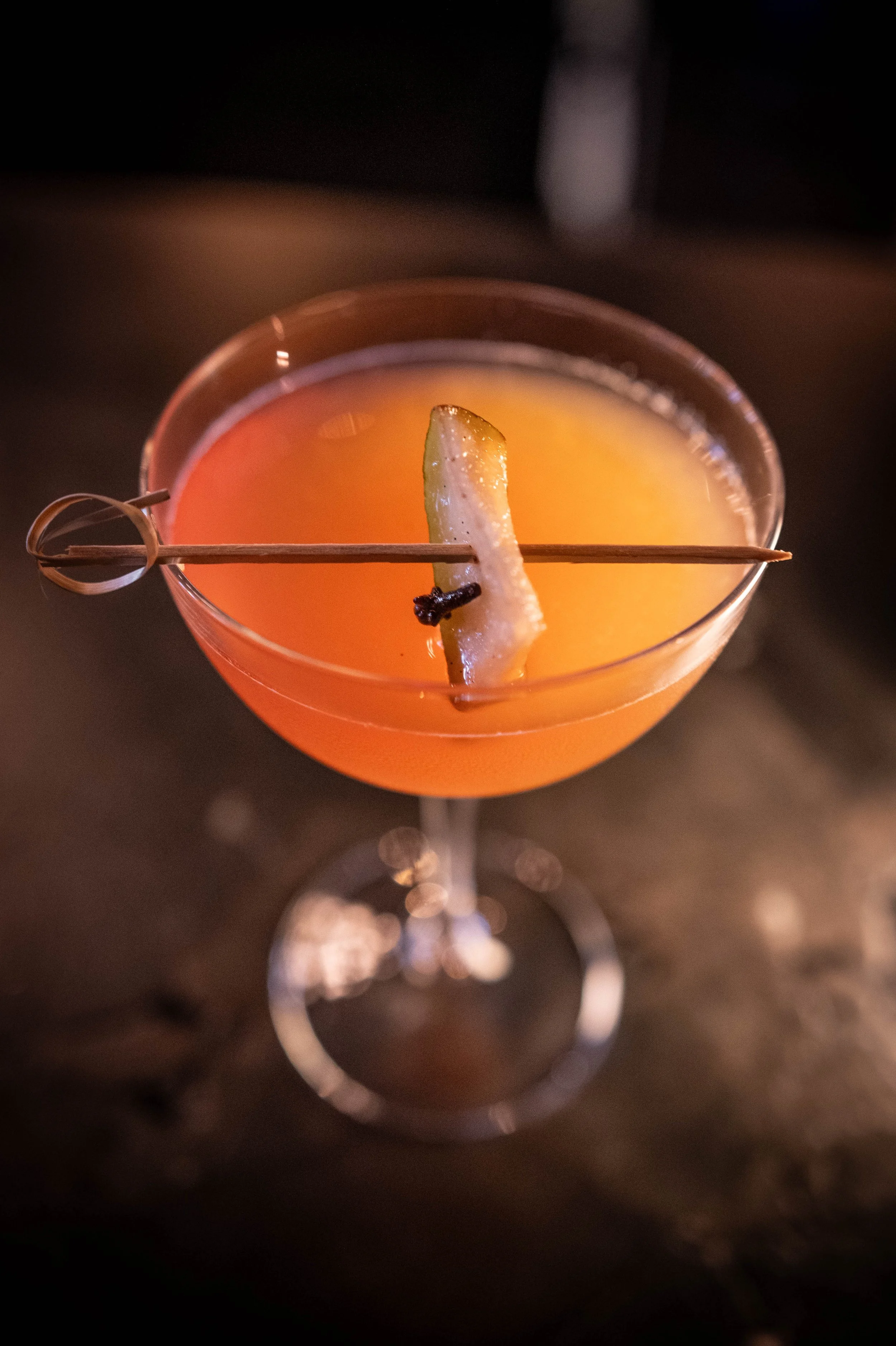 A pink cocktail in a coupe glass garnished with a lemon wedge and a small wooden skewer.