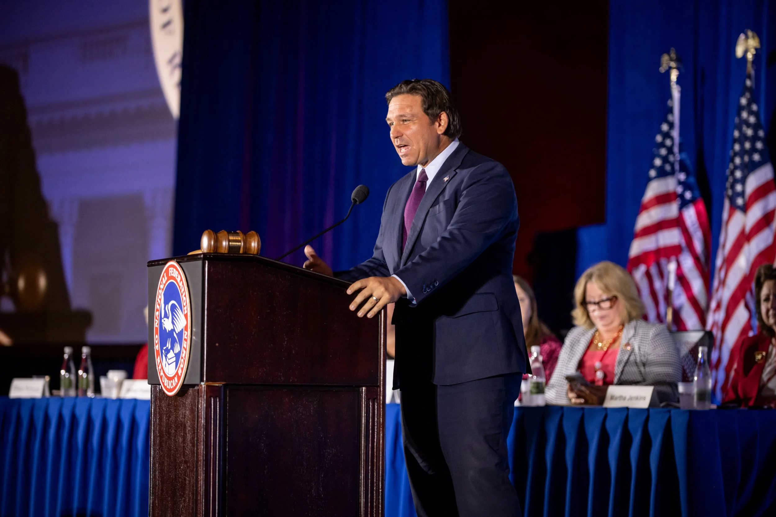 A man in a suit and purple tie is speaking at a podium with the Federal Election Commission seal, in front of a blue curtain and several seated women, some with American flags behind them, at a formal event.