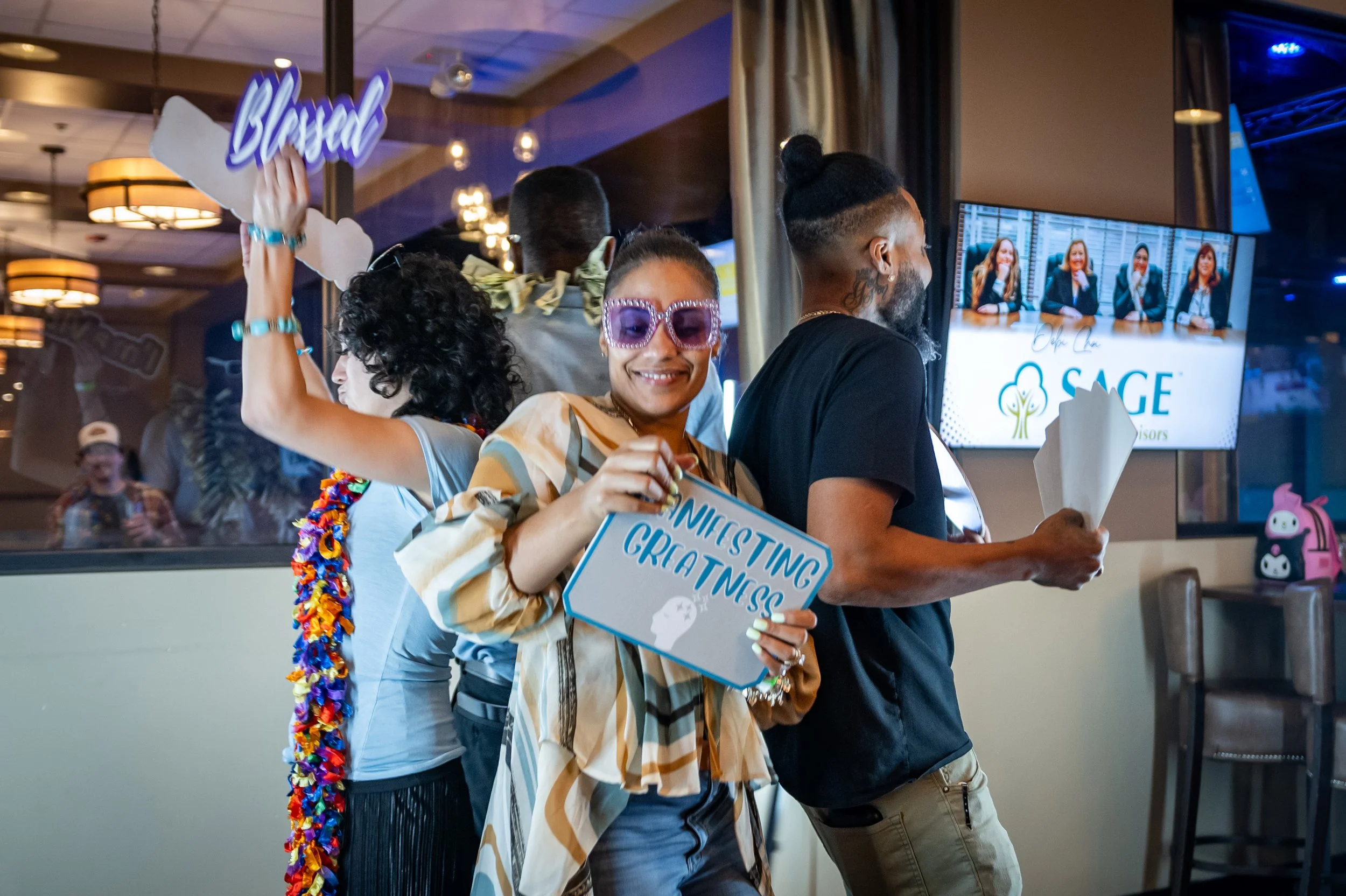 People celebrating at a graduation event indoors, with a woman holding a sign that says 'Welcoming Gratness' and smiling at the camera, others holding flowers and signs, with a TV screen in the background displaying a panel of four women.