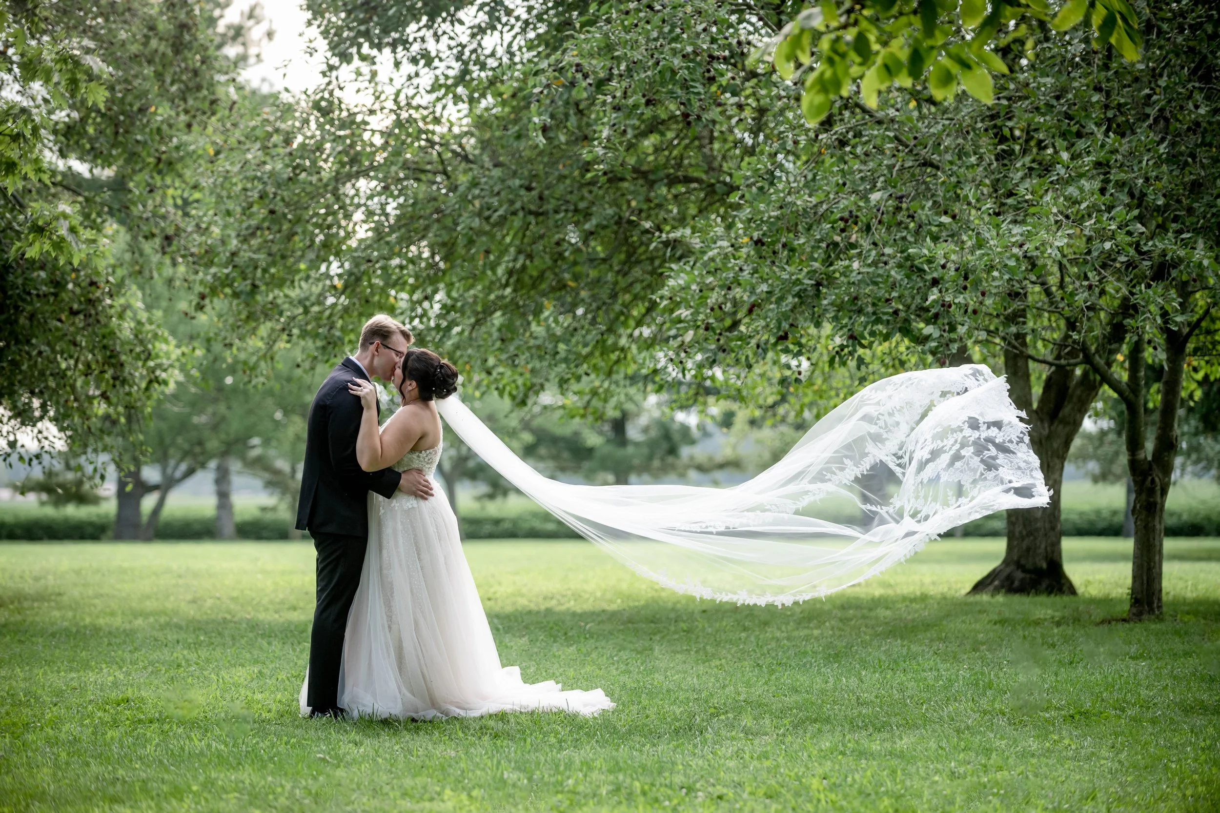 A bride and groom embracing outdoors on a grassy field, with trees in the background. The bride's long veil is flowing in the wind.