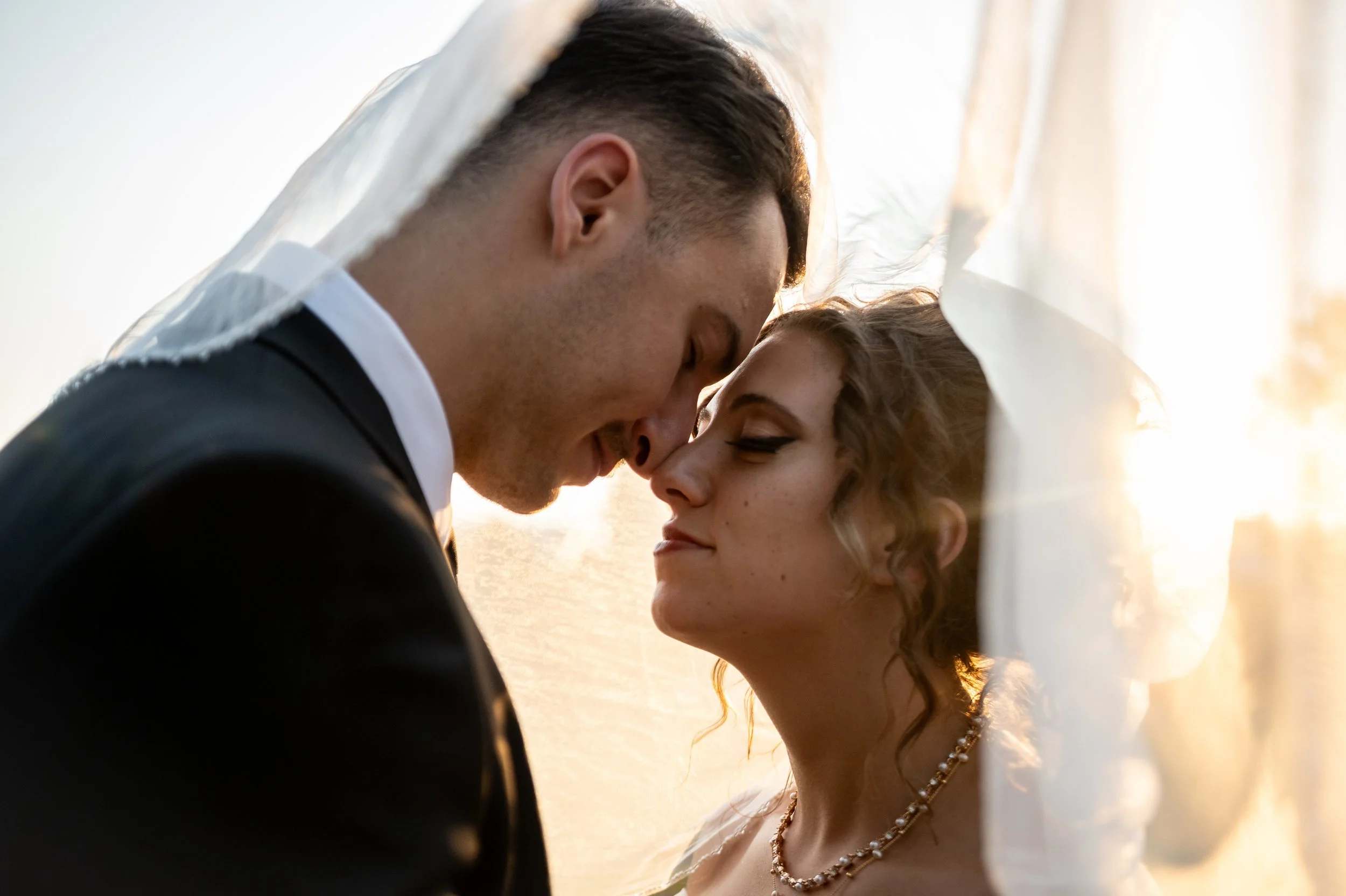 A bride and groom touching foreheads under a veil with warm sunlight in the background.
