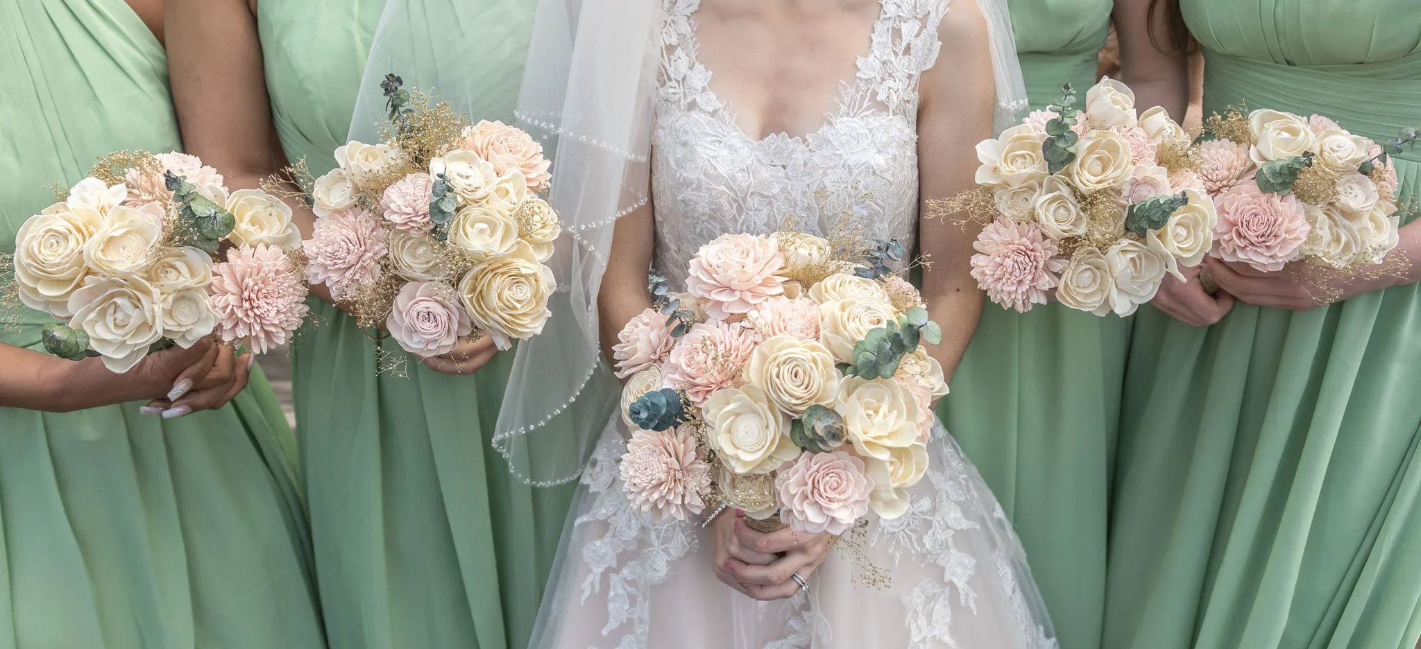 Bride in white lace wedding dress holding a bouquet of pastel pink and white roses, light pink dahlias, and eucalyptus, flanked by bridesmaids in light green dresses holding similar bouquets.