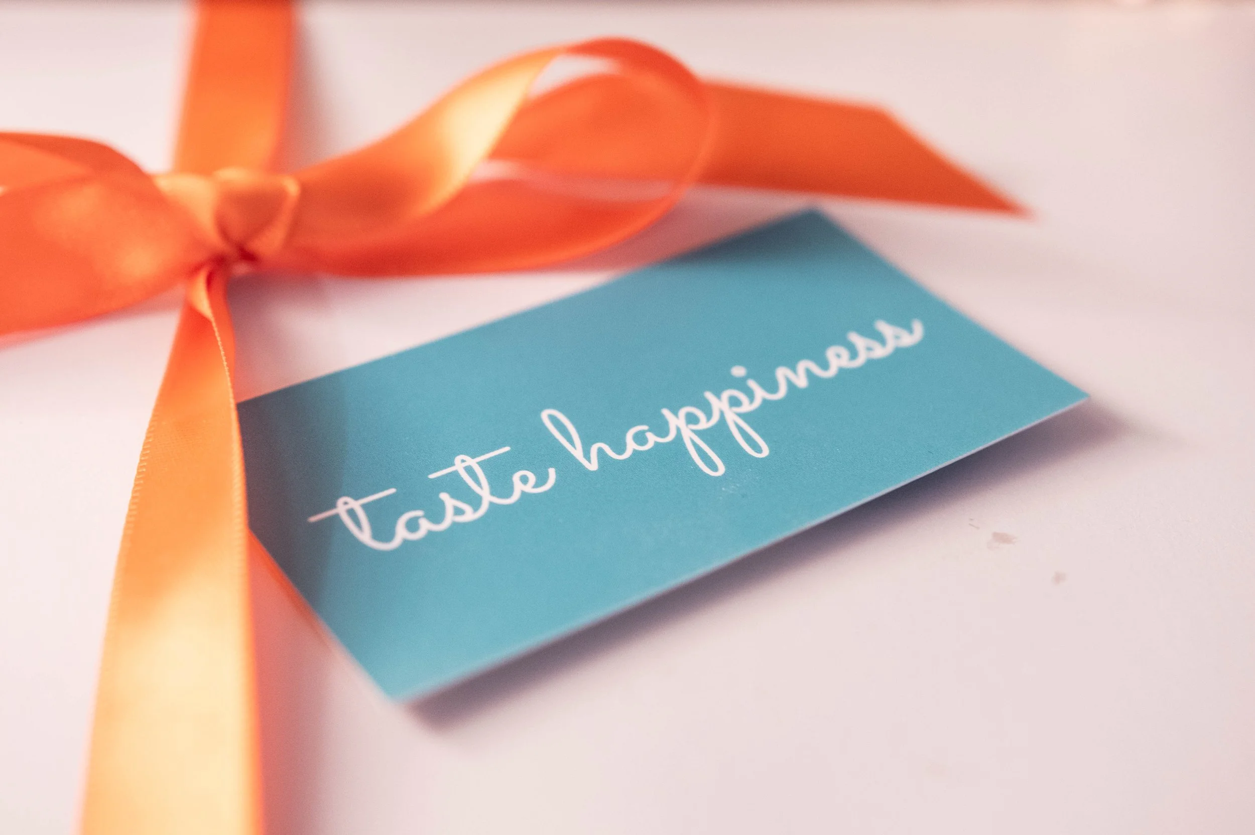 A blue card with white cursive writing reading 'taste happiness', attached to a peach-colored satin ribbon tied in a bow.