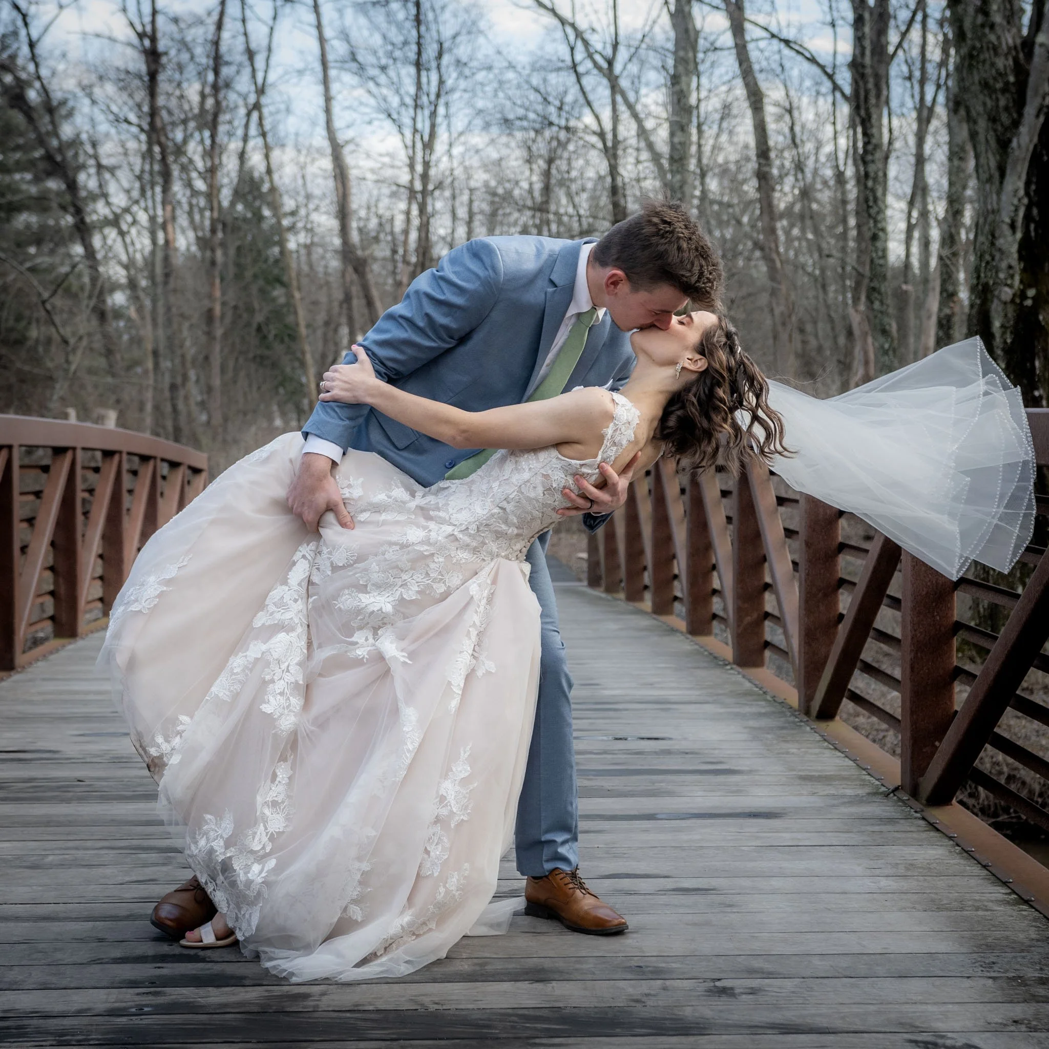 A groom in a blue suit and a bride in a wedding dress sharing a kiss on a wooden bridge in a forested area.