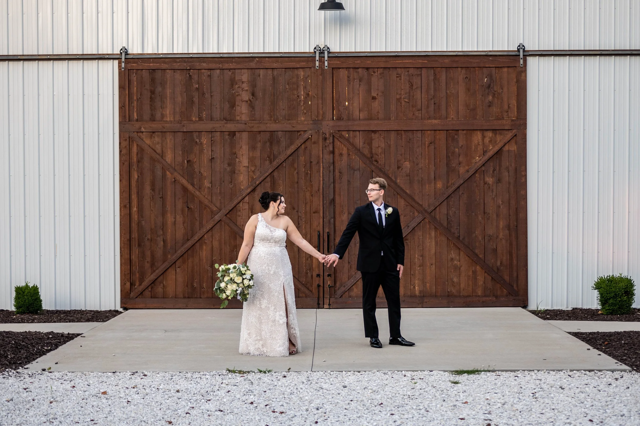 A bride and groom holding hands in front of a large wooden barn door. The bride is in a lace wedding gown holding a bouquet, and the groom is in a black suit with a white boutonniere.