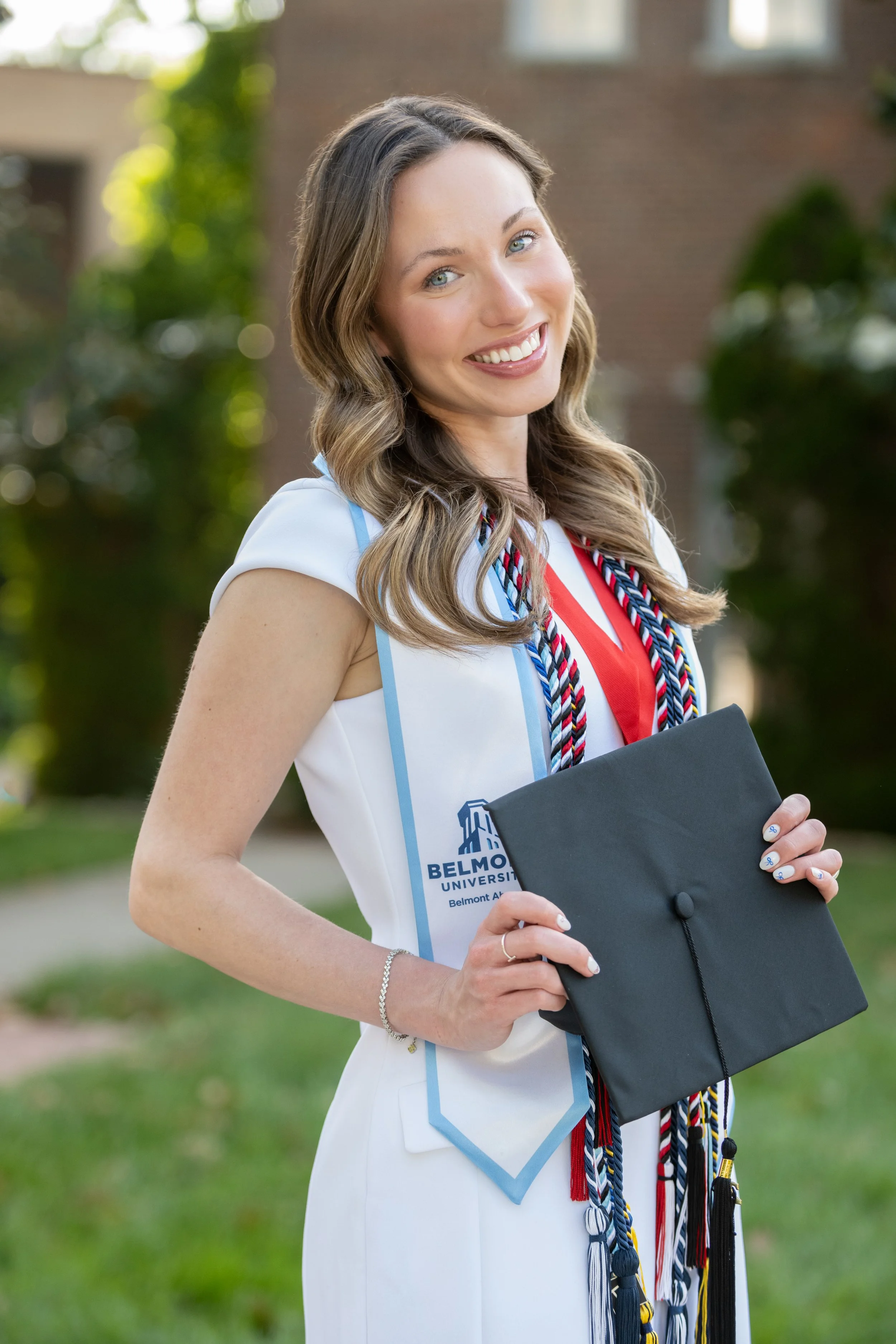 A young woman wearing a white graduate gown and holding a black graduation cap, standing outdoors with greenery in the background, smiling and looking at the camera.