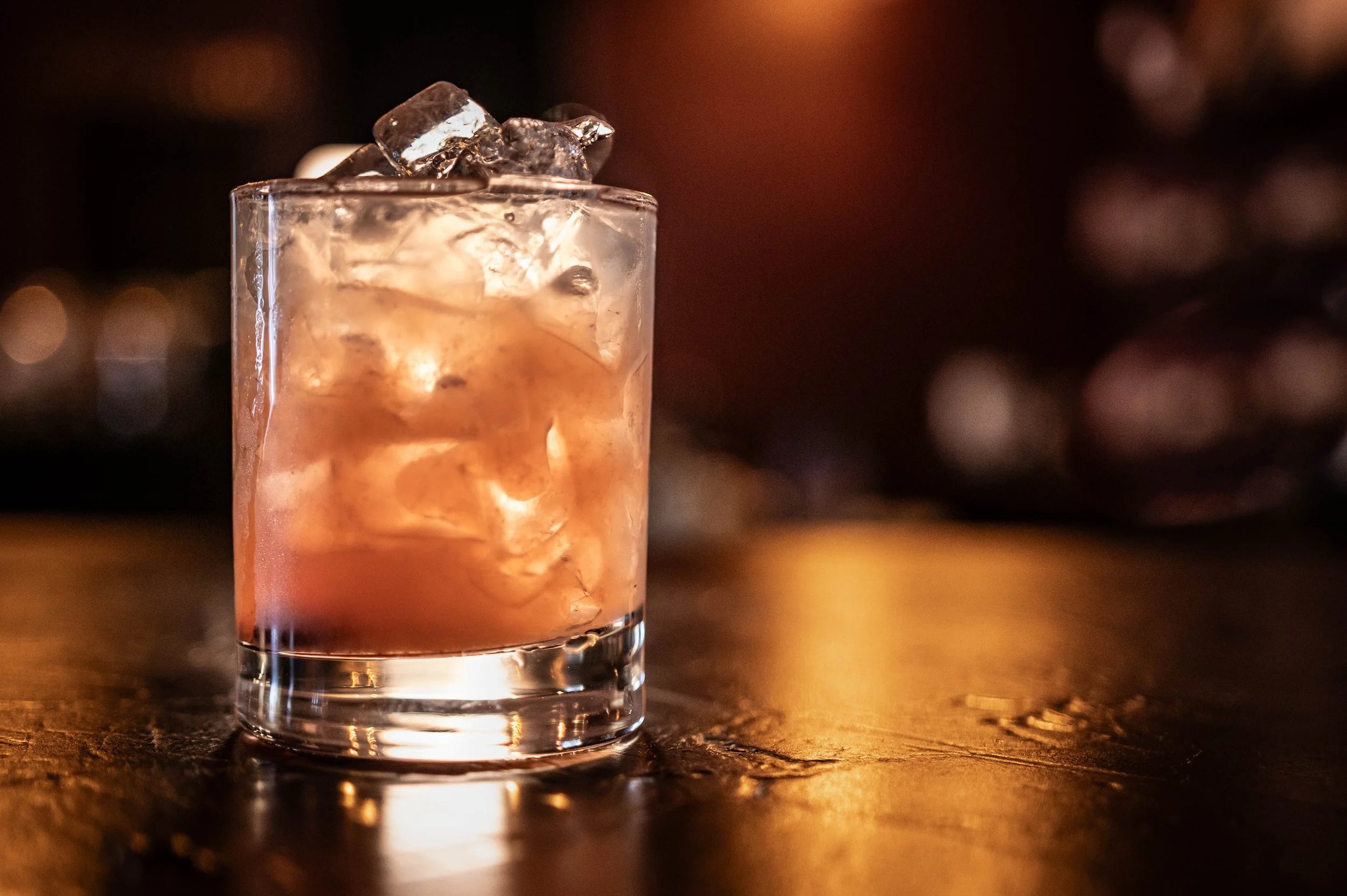 A glass filled with a pink cocktail and ice cubes, placed on a wooden surface with blurred warm lighting in the background.