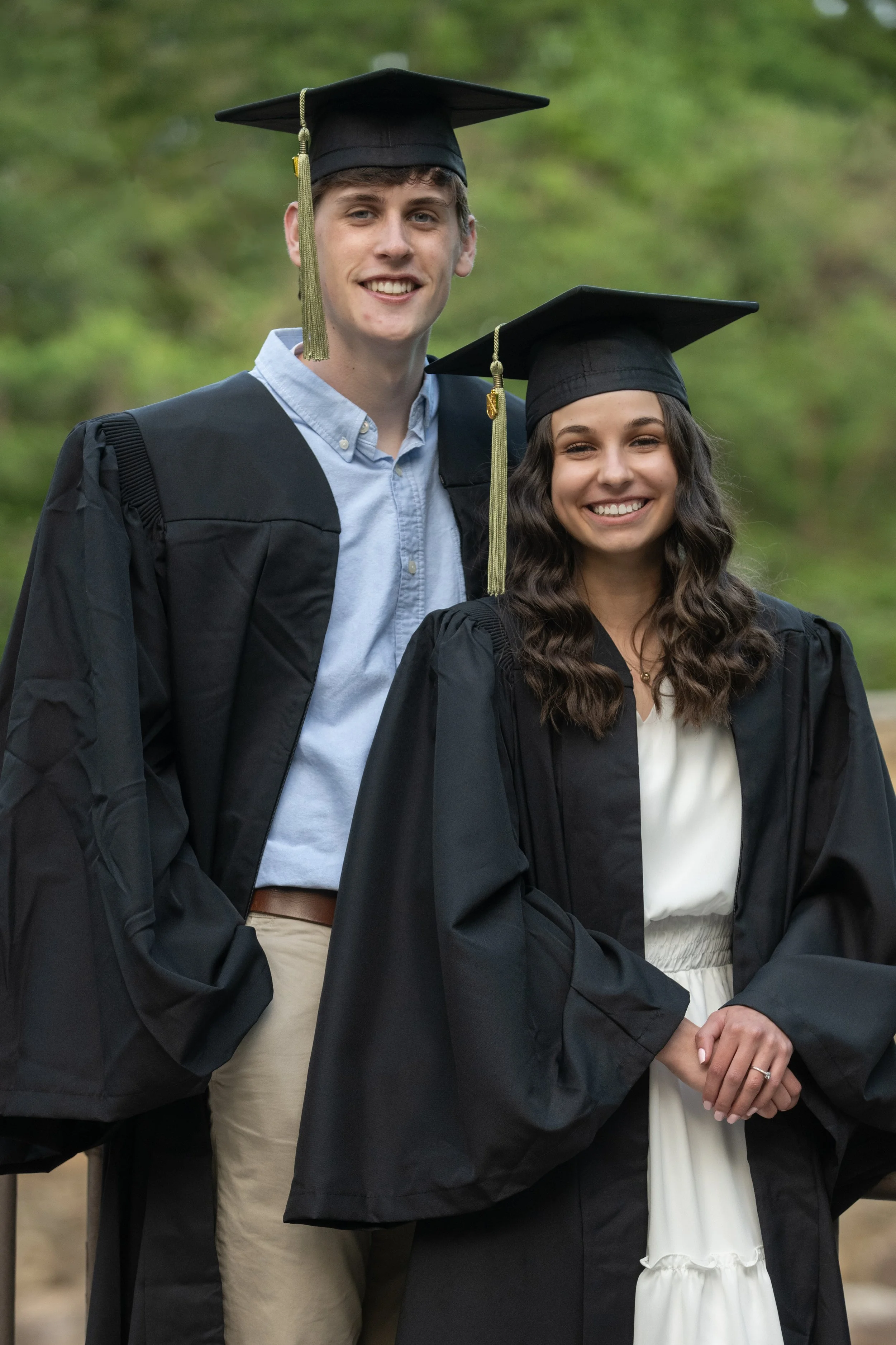 Two young graduates, a male and a female, wearing black caps and gowns, smiling outdoors during graduation ceremony.