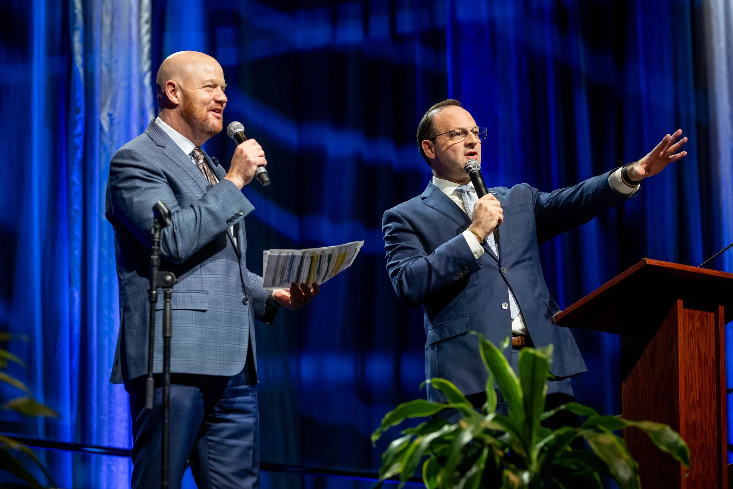 Two men in suits speaking into microphones on stage during an event, with blue curtains and a wooden podium, and a green plant in the foreground.