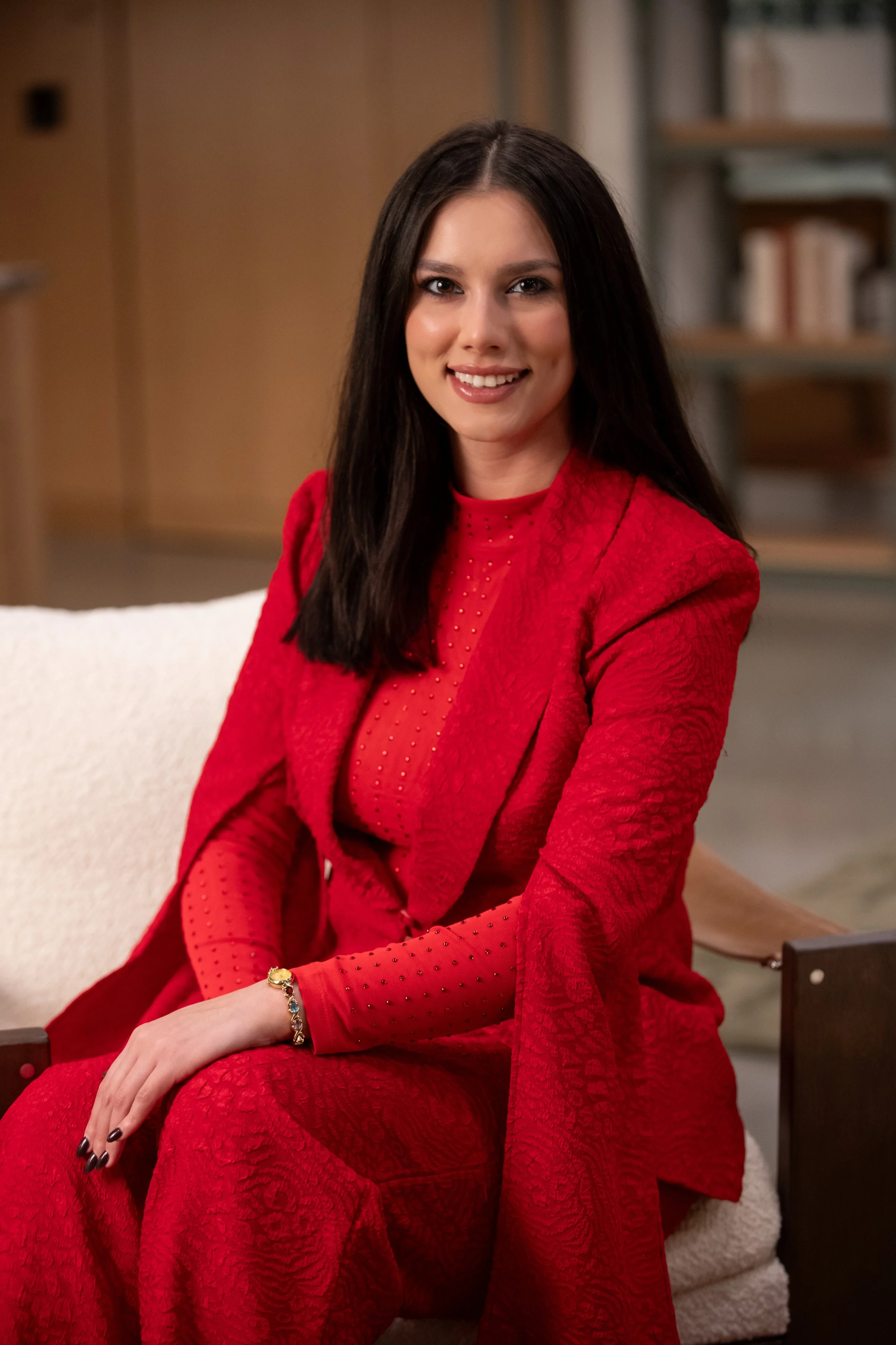 A woman with long dark hair sitting on a beige couch, smiling and wearing a red patterned suit with a red sheer top, accessorized with a gold and gemstone bracelet. In the background, a bookshelf is visible.