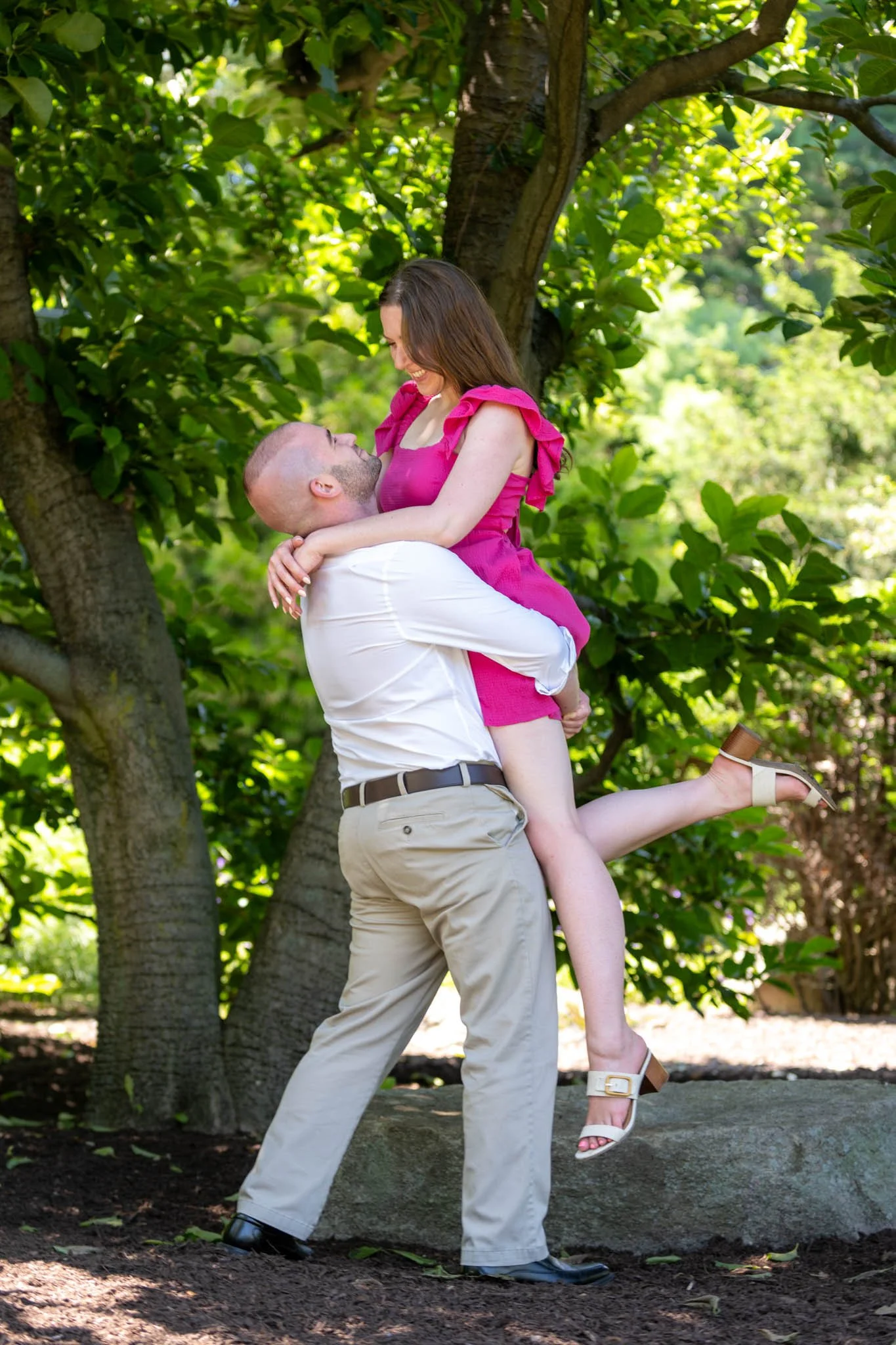 A man lifting a woman outdoors in a park with lush green trees, both smiling and looking at each other, with the woman wearing a pink dress and white heels.