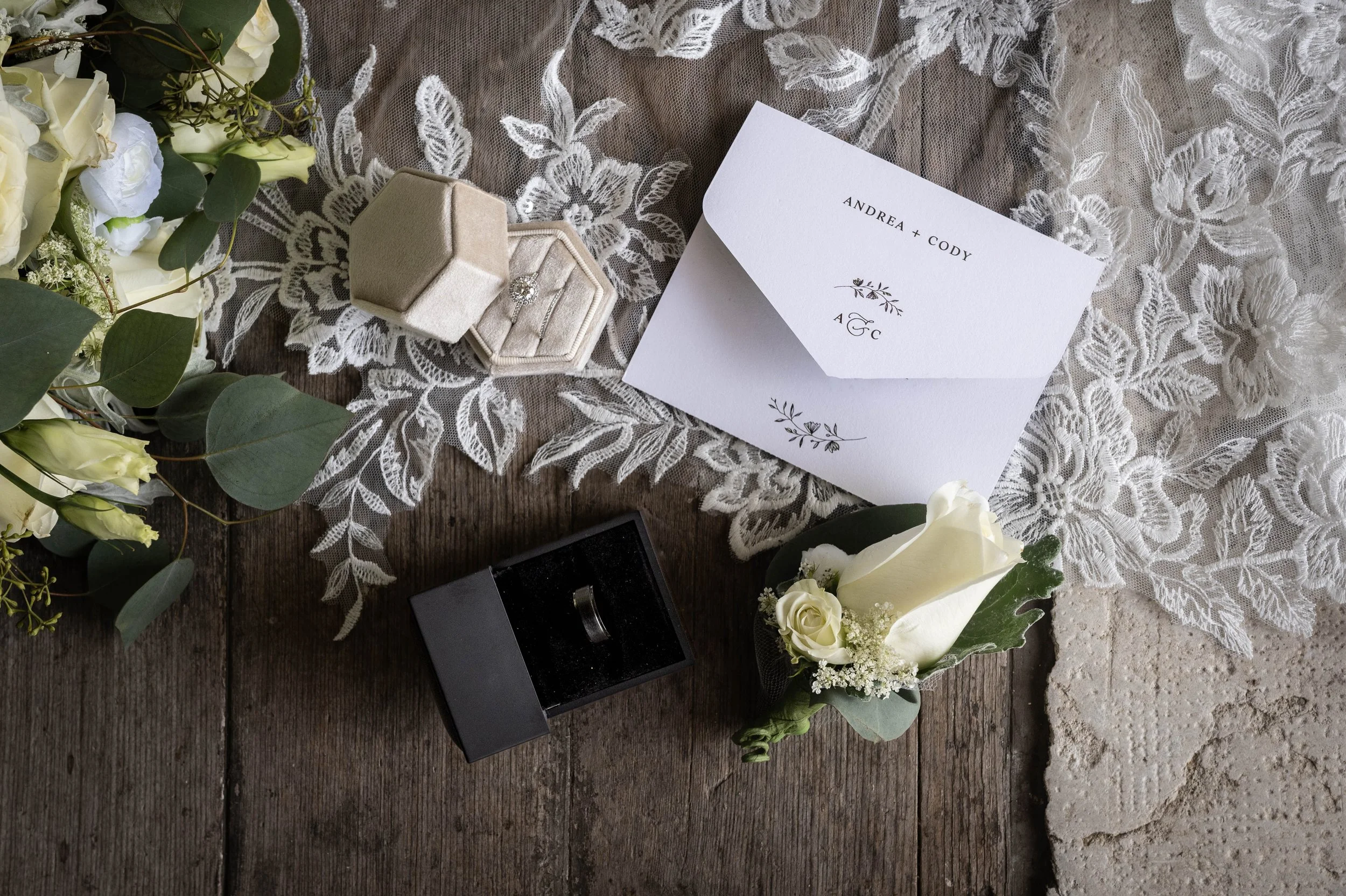 Wedding invitation, engagement ring box, and white floral arrangement on lace and wooden surface.