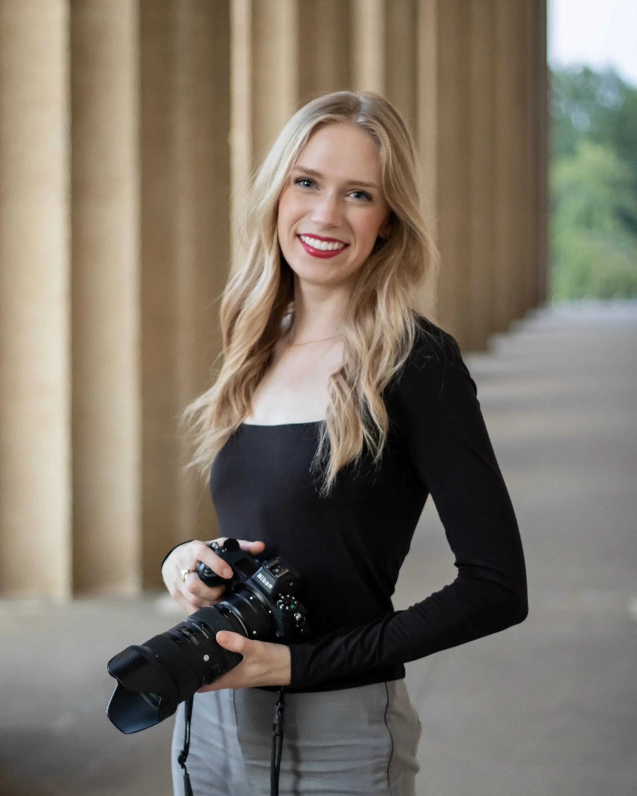A young woman with long blonde hair and red lipstick holding a professional camera in an outdoor location with large columns, greenery, and a walkway.