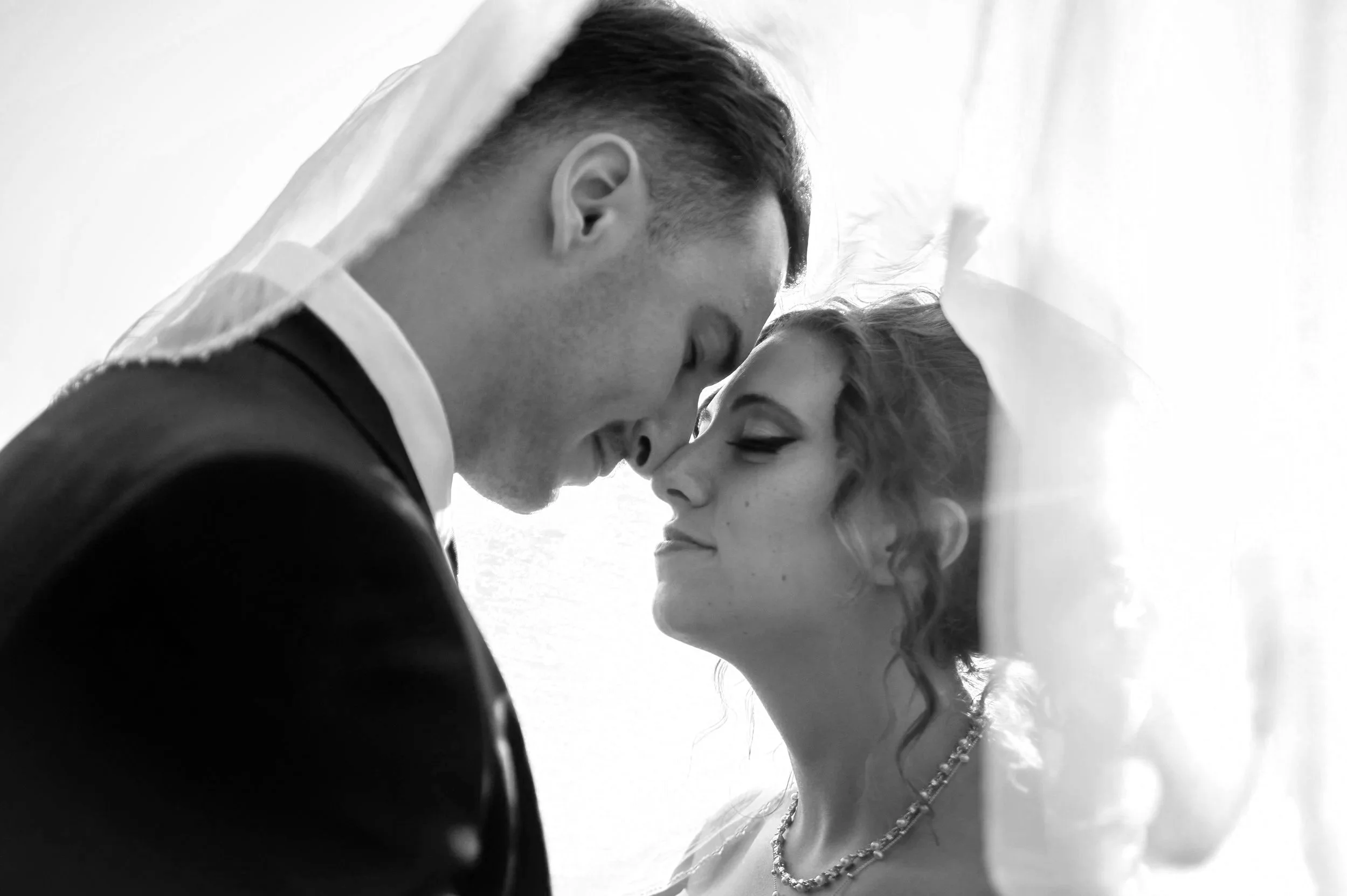 A couple, a man and woman, touch foreheads with their eyes closed, sharing an intimate moment. The woman wears a pearl necklace and has styled hair, while the man wears a tuxedo with a veil over his head. The scene is black and white.