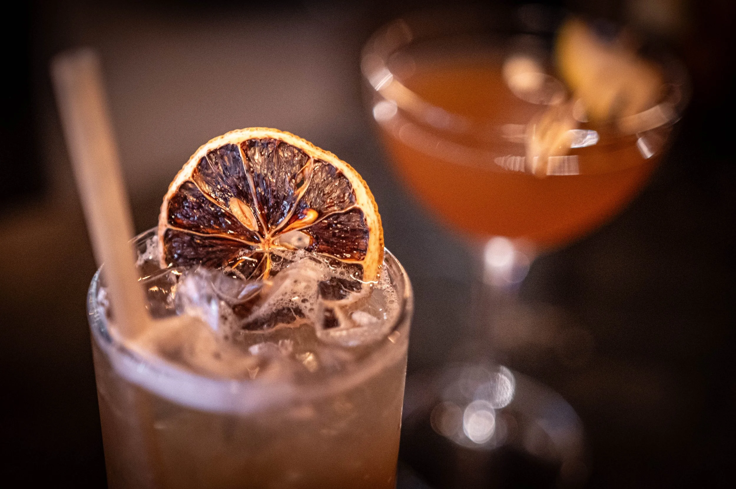 A beverage with ice and a dried lemon slice garnish in a glass with a straw, and a cocktail in a coupe glass in the background.