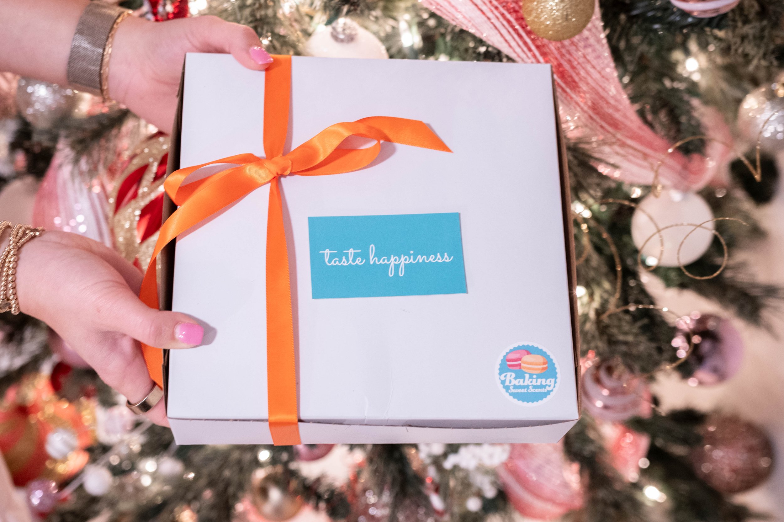 Person holding a white gift box with orange ribbon, decorated with a blue label that says 'taste happiness' and a small sticker with macarons and the words 'Baking Sweet Scents', in front of a decorated Christmas tree