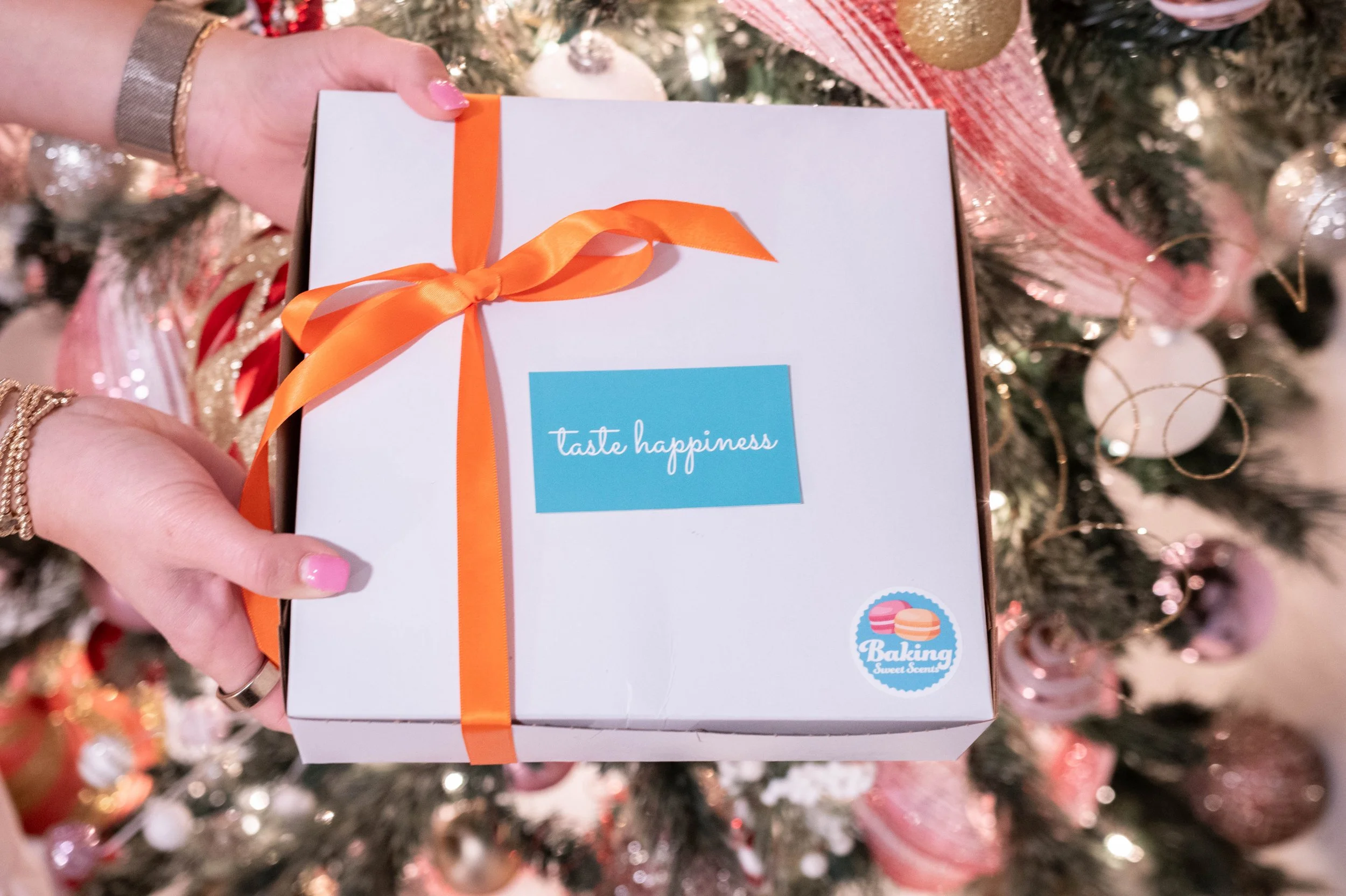 Person holding a white gift box with orange ribbon, decorated with a blue label that says 'taste happiness' and a small sticker with macarons and the words 'Baking Sweet Scents', in front of a decorated Christmas tree