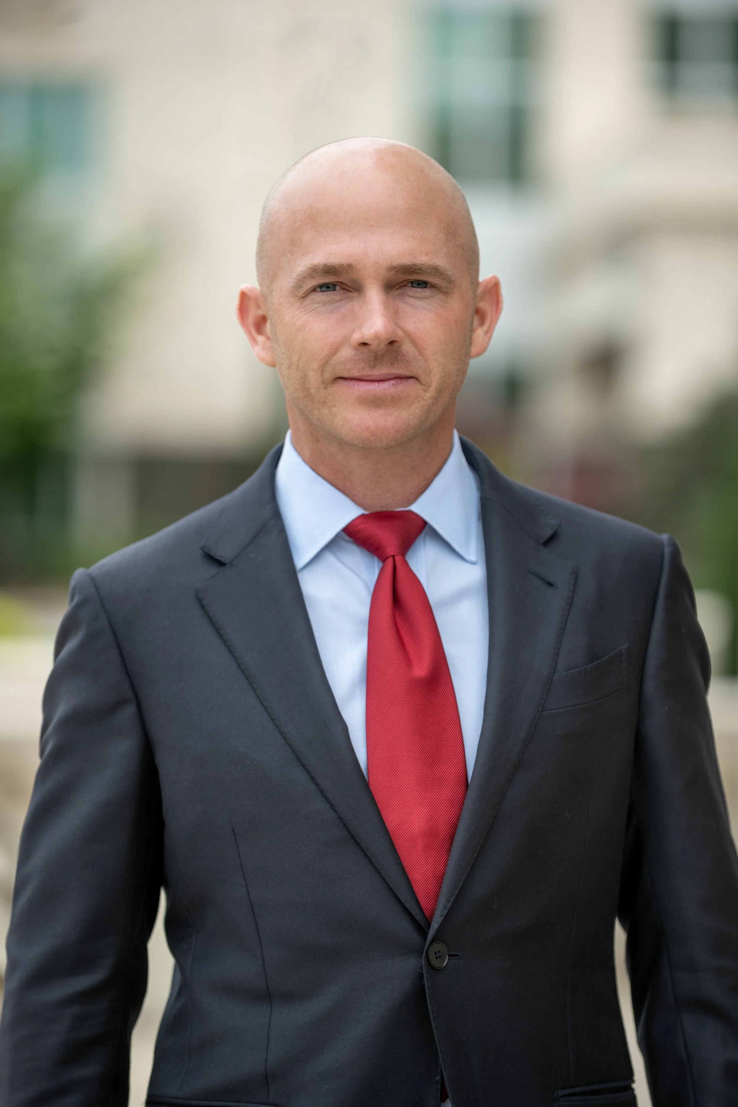 A bald man wearing a dark suit, light blue shirt, and red tie standing outdoors in front of a blurred background.