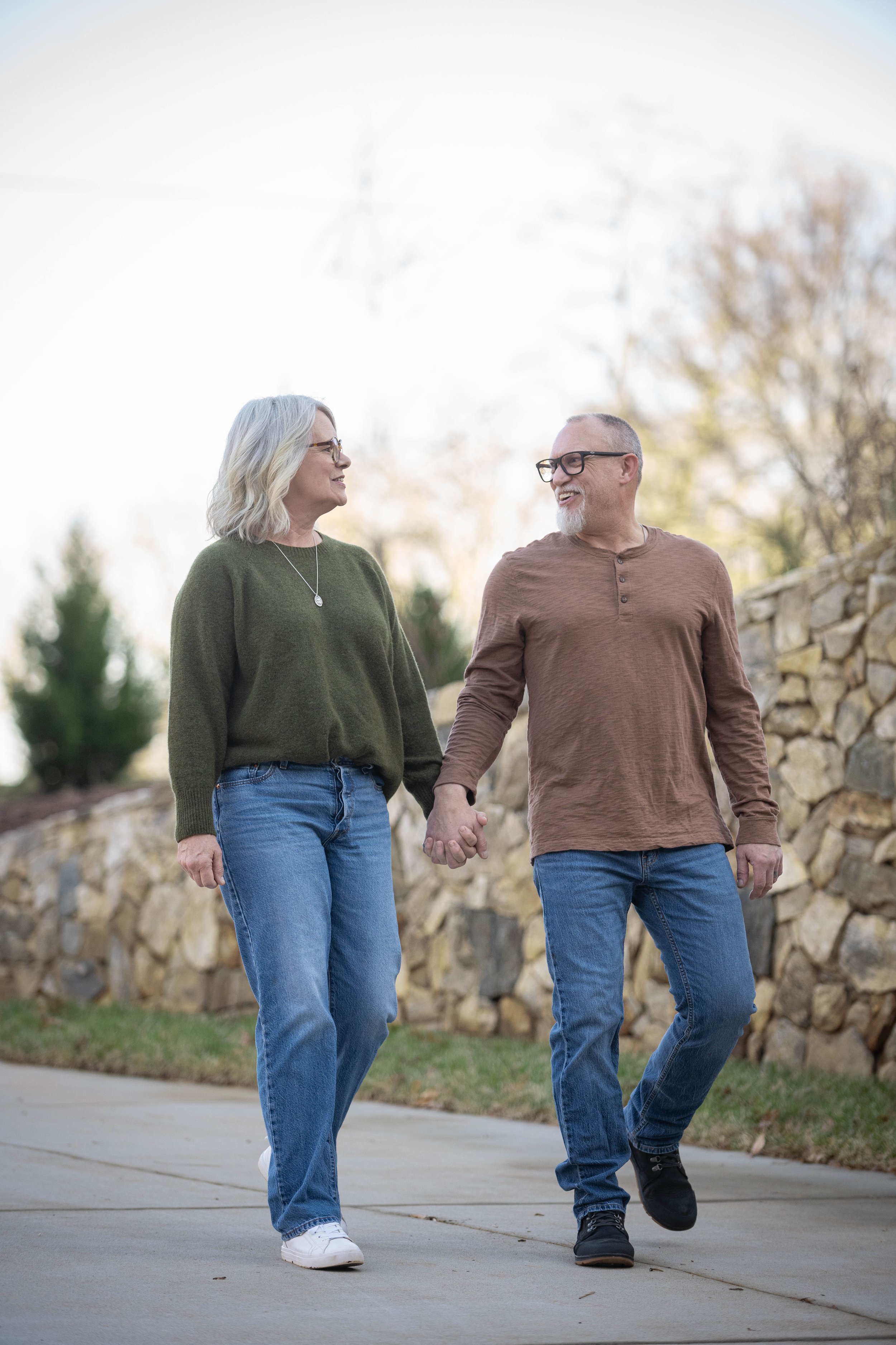 A middle-aged couple holding hands and walking outdoors on a sidewalk, smiling, with a stone wall and trees in the background.
