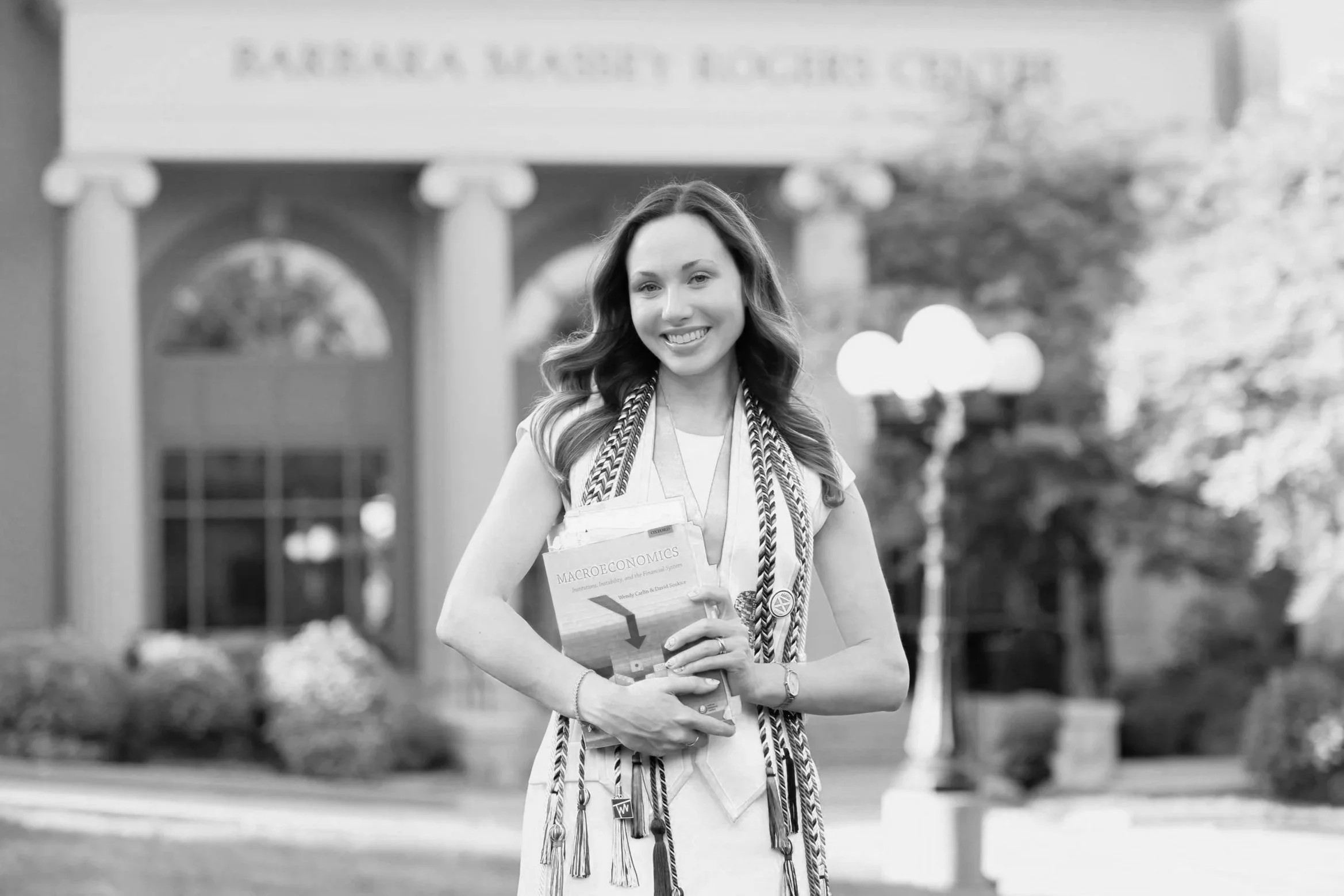 A smiling woman holding a book titled 'Macroeconomics' standing outside a building with columns and a sign above the entrance.