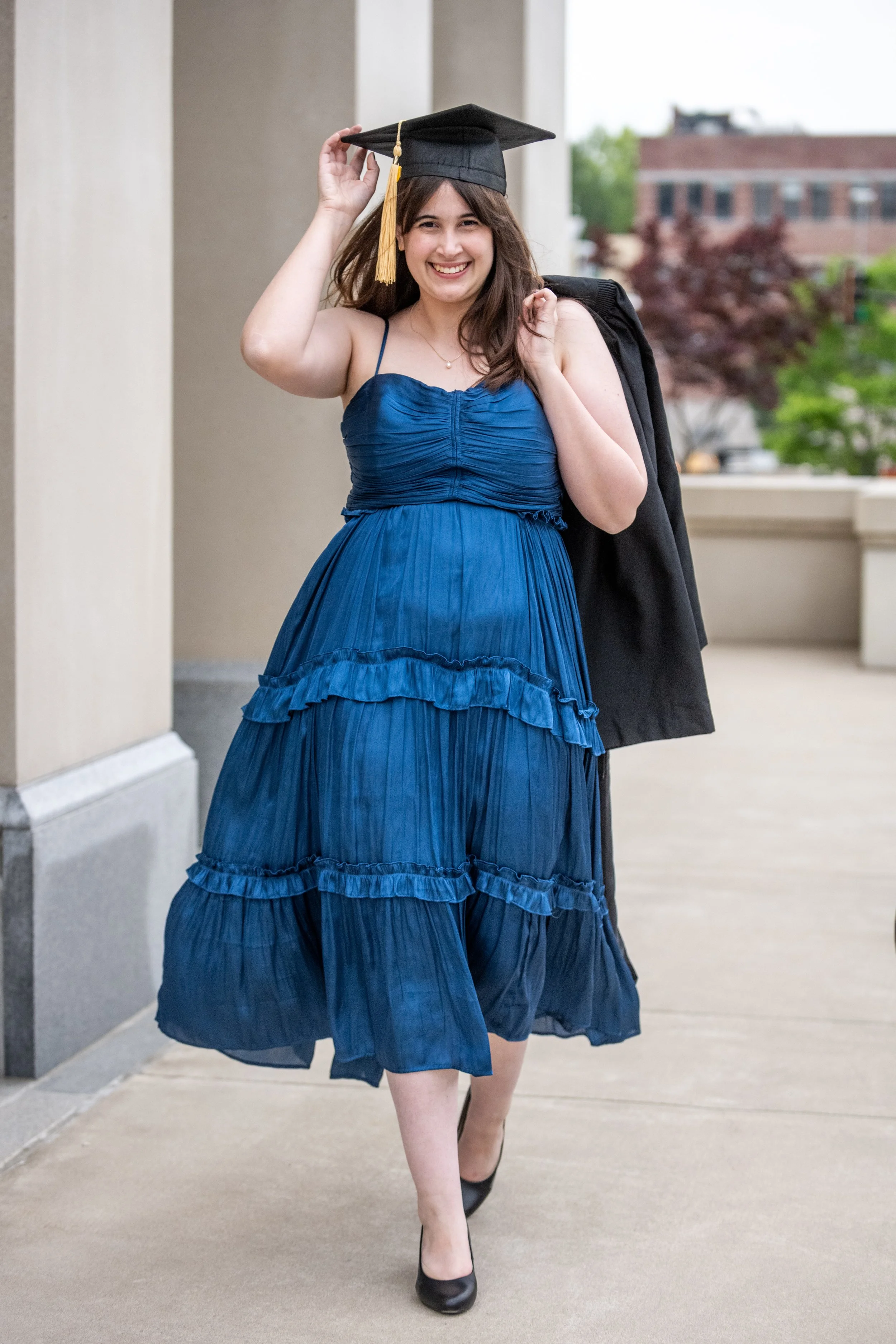 A young woman in a blue dress and black heels celebrating graduation with a cap and gown over her shoulder, smiling and walking outdoors.