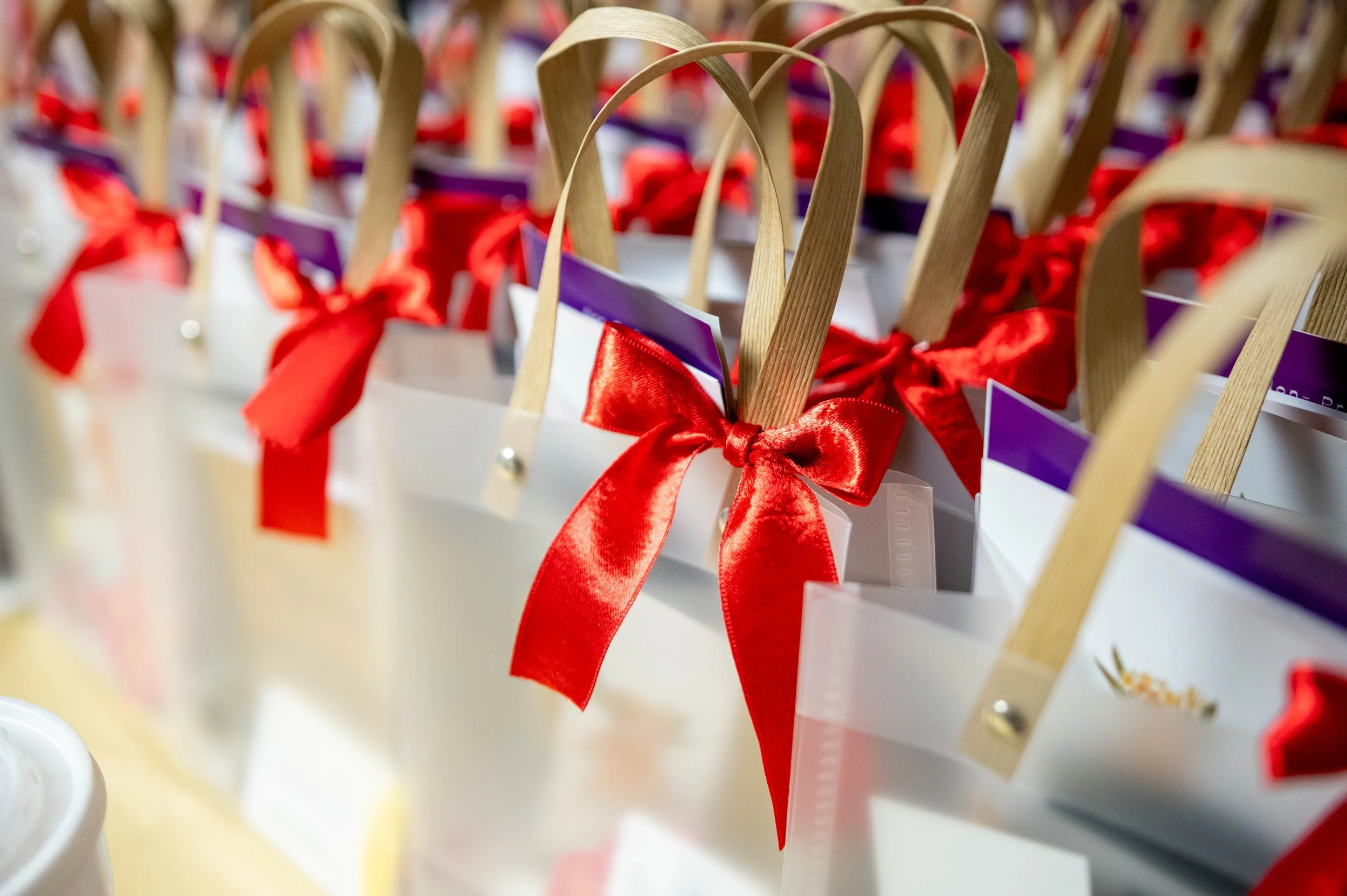 Close-up of white gift bags with red satin bows and wooden handles, arranged in a row.