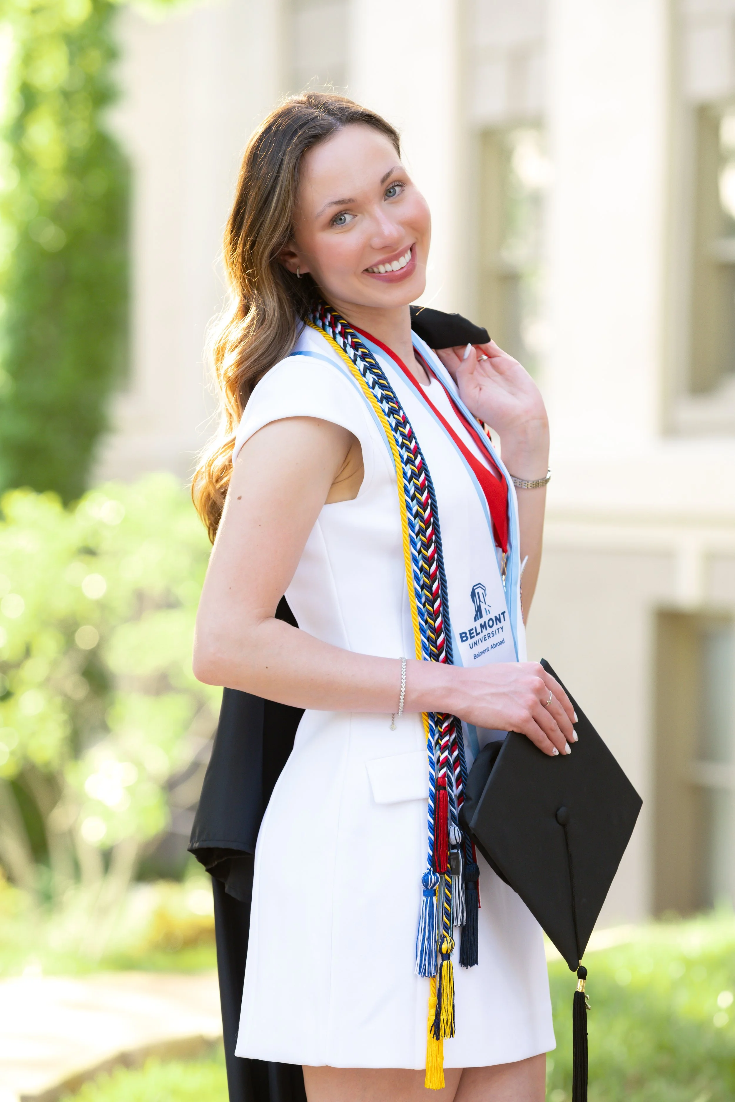 A smiling young woman in a white dress holding a graduation cap outdoors, wearing multiple honor cords, with a Belmont University badge around her neck.