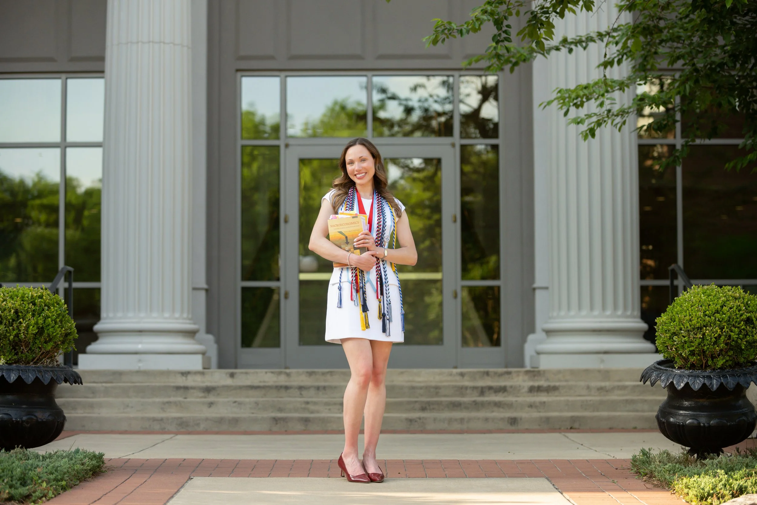 A young woman in a white dress and high heels stands on steps in front of a building with large glass doors, holding a diploma and a book, smiling.