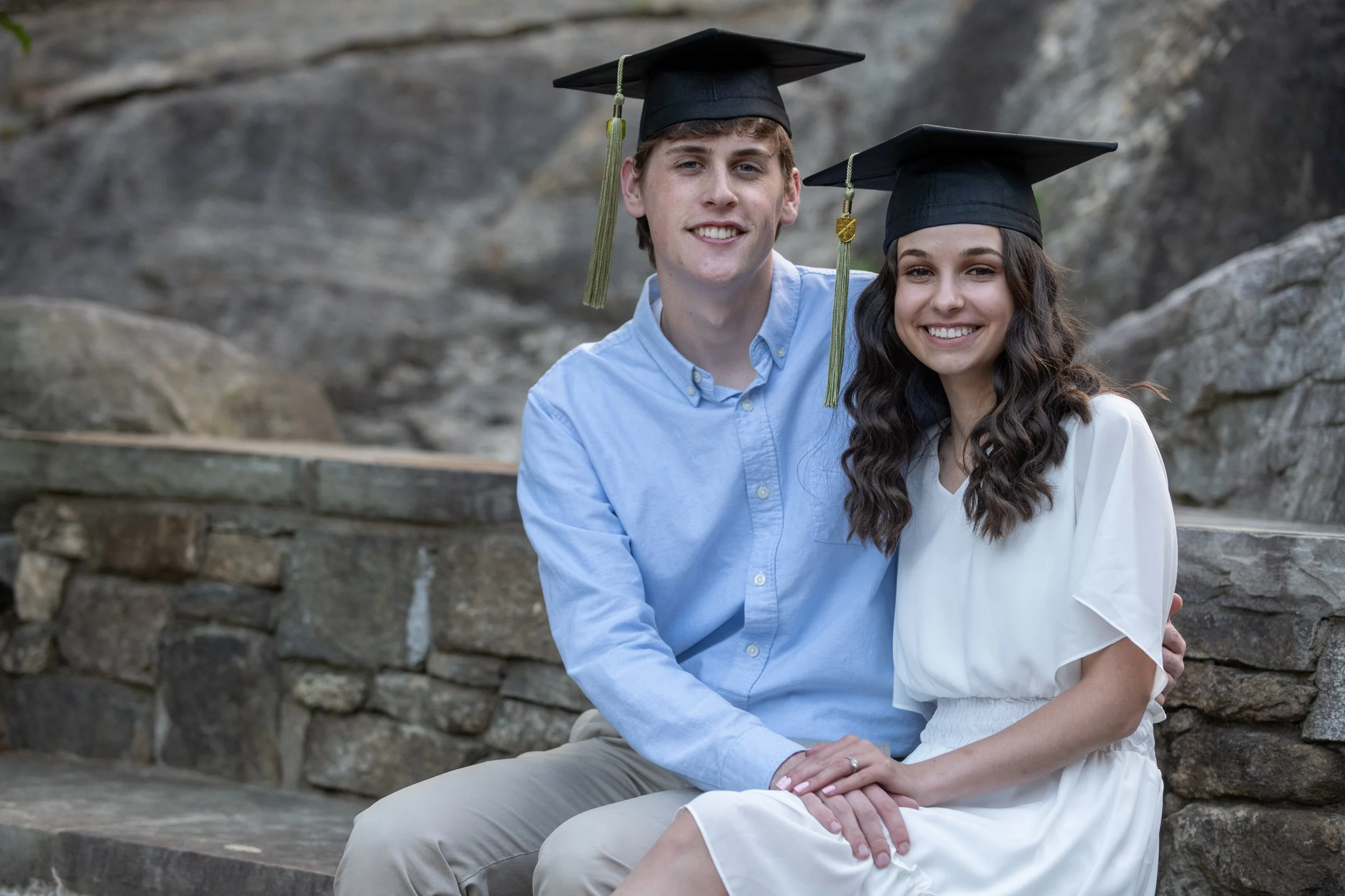 Young man and woman in graduation caps sitting together outdoors, smiling at the camera.