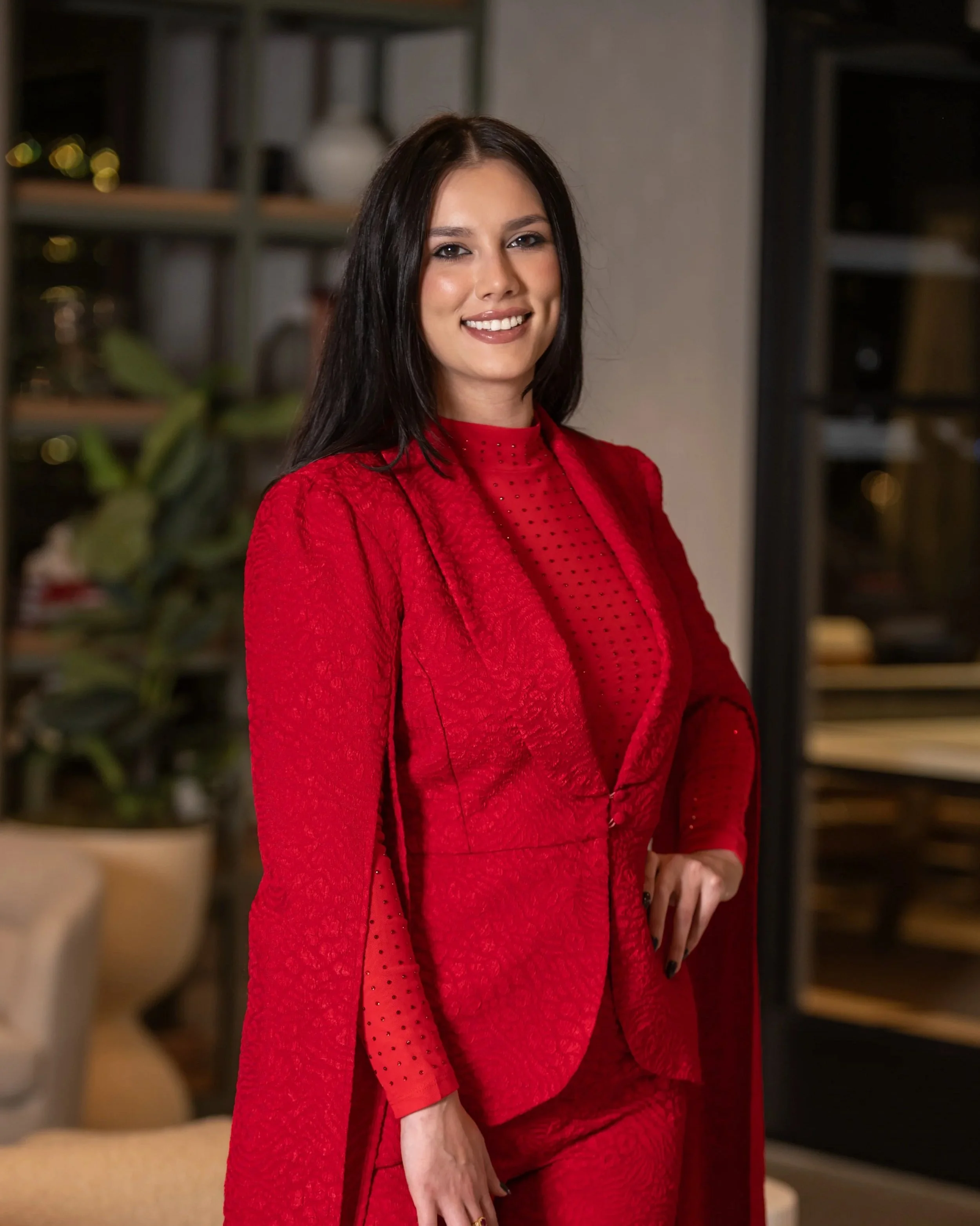 A woman wearing a red textured suit and a red top, smiling, standing in a room with blurred shelves and indoor plants in the background.