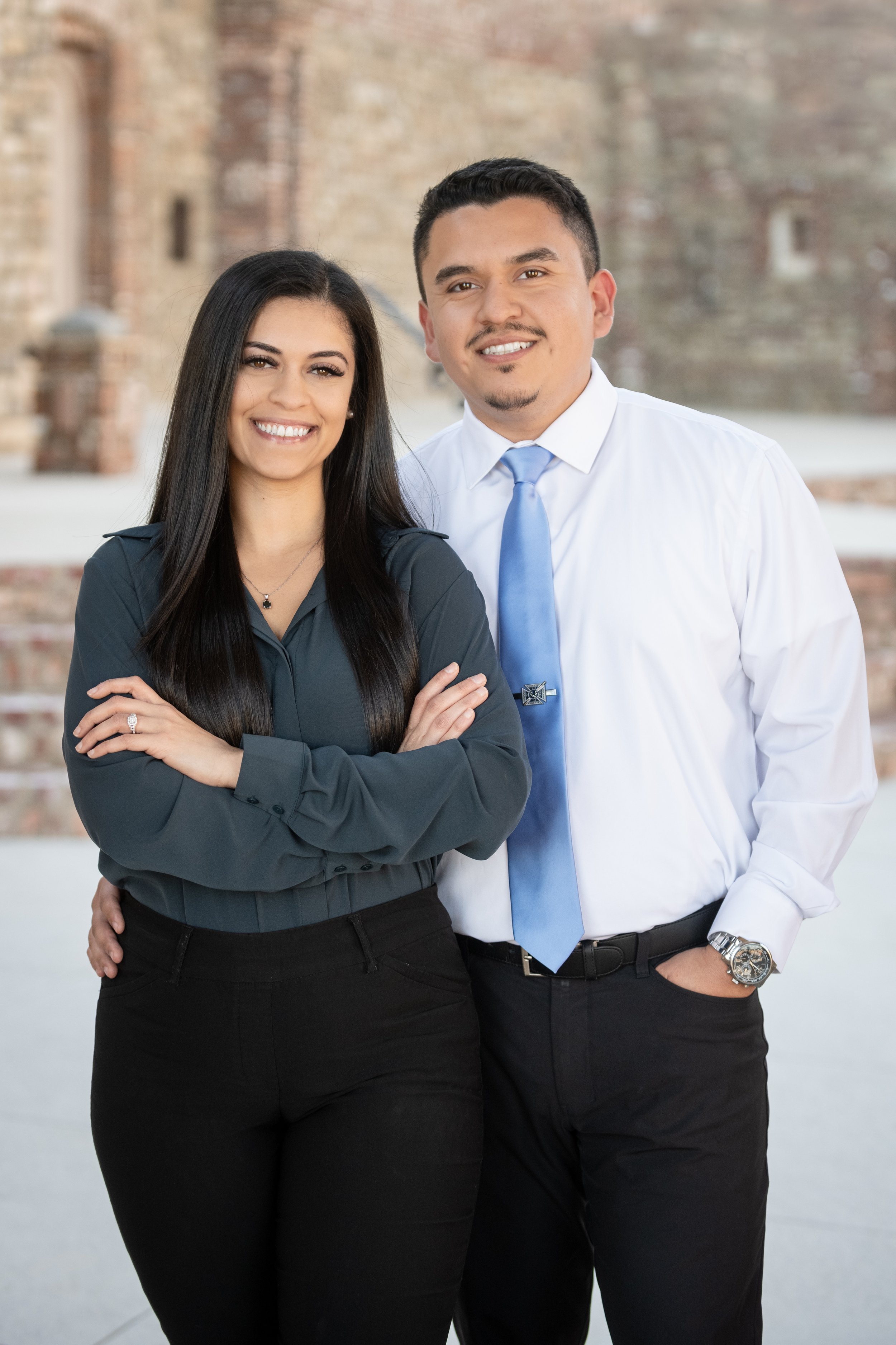 A smiling woman in a dark blouse with long dark hair standing next to a smiling man in a white shirt, blue tie, and dark pants outside in front of a brick building.