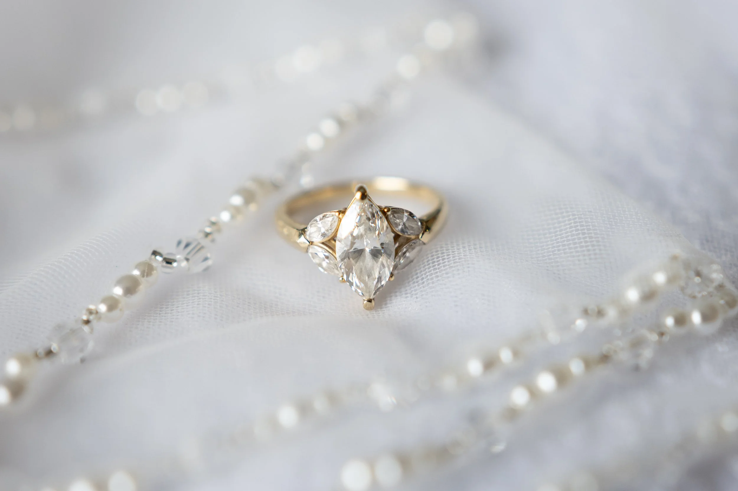 A gold ring with a large marquise-shaped diamond and two smaller pear-shaped diamonds on either side, resting on white fabric with a pearl necklace draped around it.