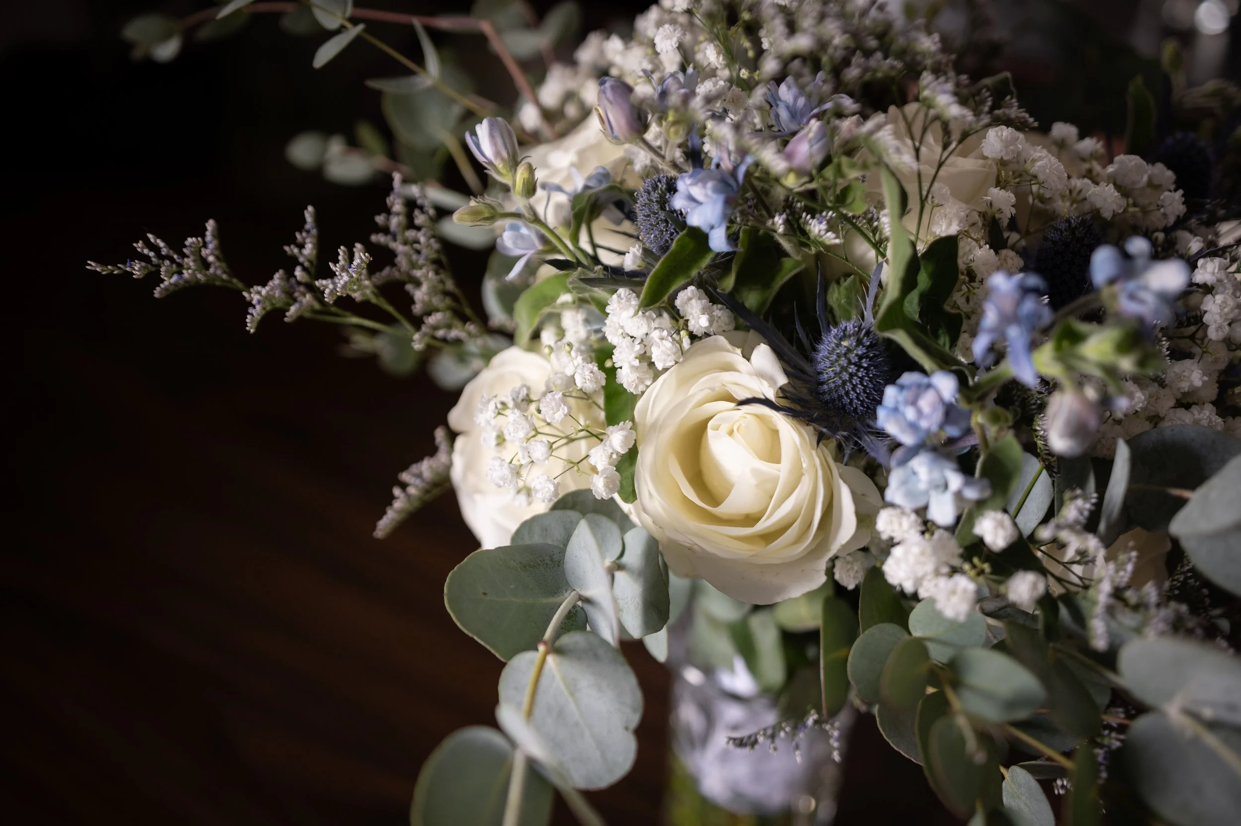 A wedding bouquet with white roses, blue thistles, baby's breath, green eucalyptus leaves, and other small flowers.