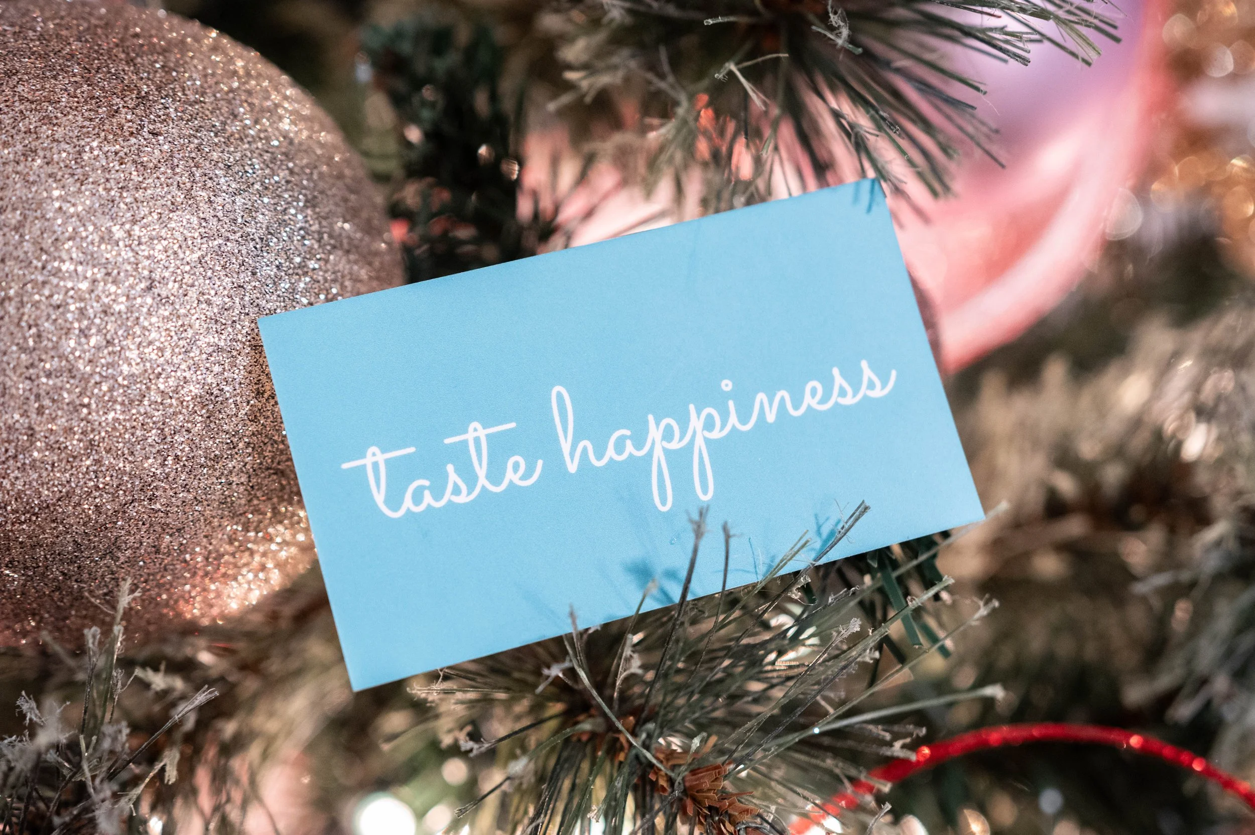 Close-up of a Christmas tree decorated with a large glittery ornament and a pink ribbon, with a blue card reading 'taste happiness' attached to the branches.