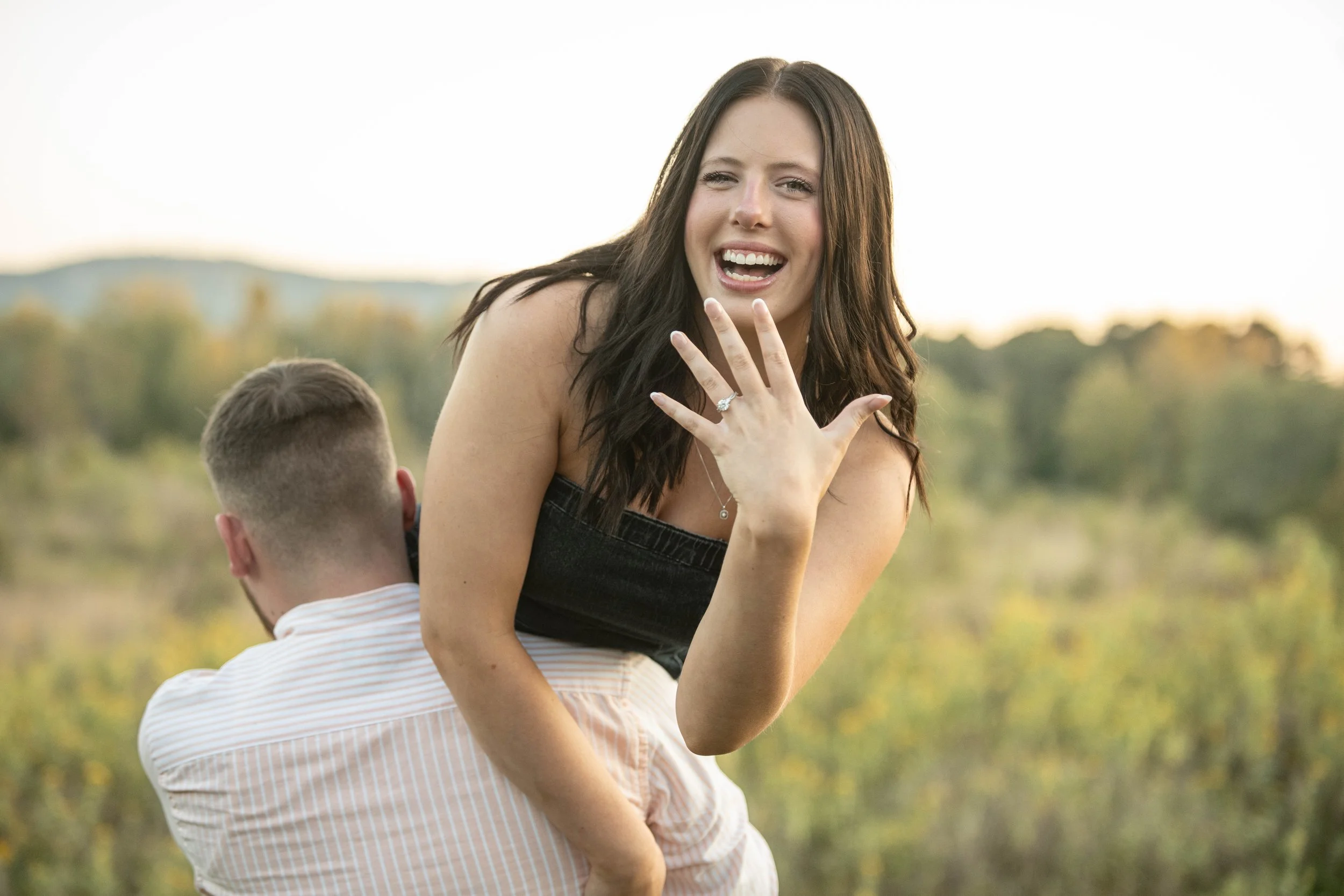 A woman with long dark hair, wearing a black strapless top, shows her engagement ring while sitting on a man's back outdoors with a blurred landscape of trees and hills in the background.