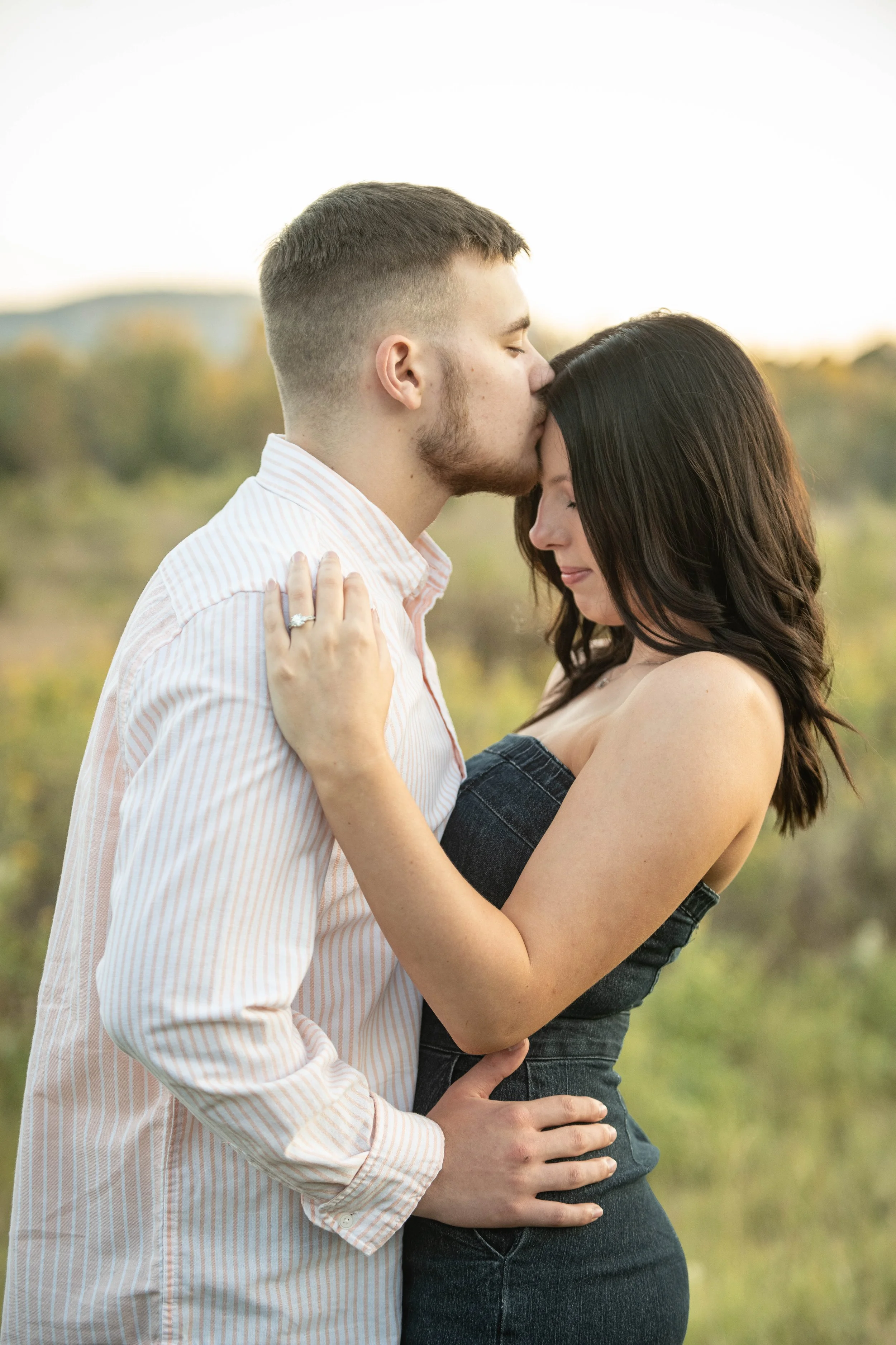 A man and woman embracing outdoors, with the man kissing the woman's forehead, and both holding each other gently. The woman has dark hair and is wearing a strapless denim dress, while the man has short hair and is wearing a light-colored, striped sh