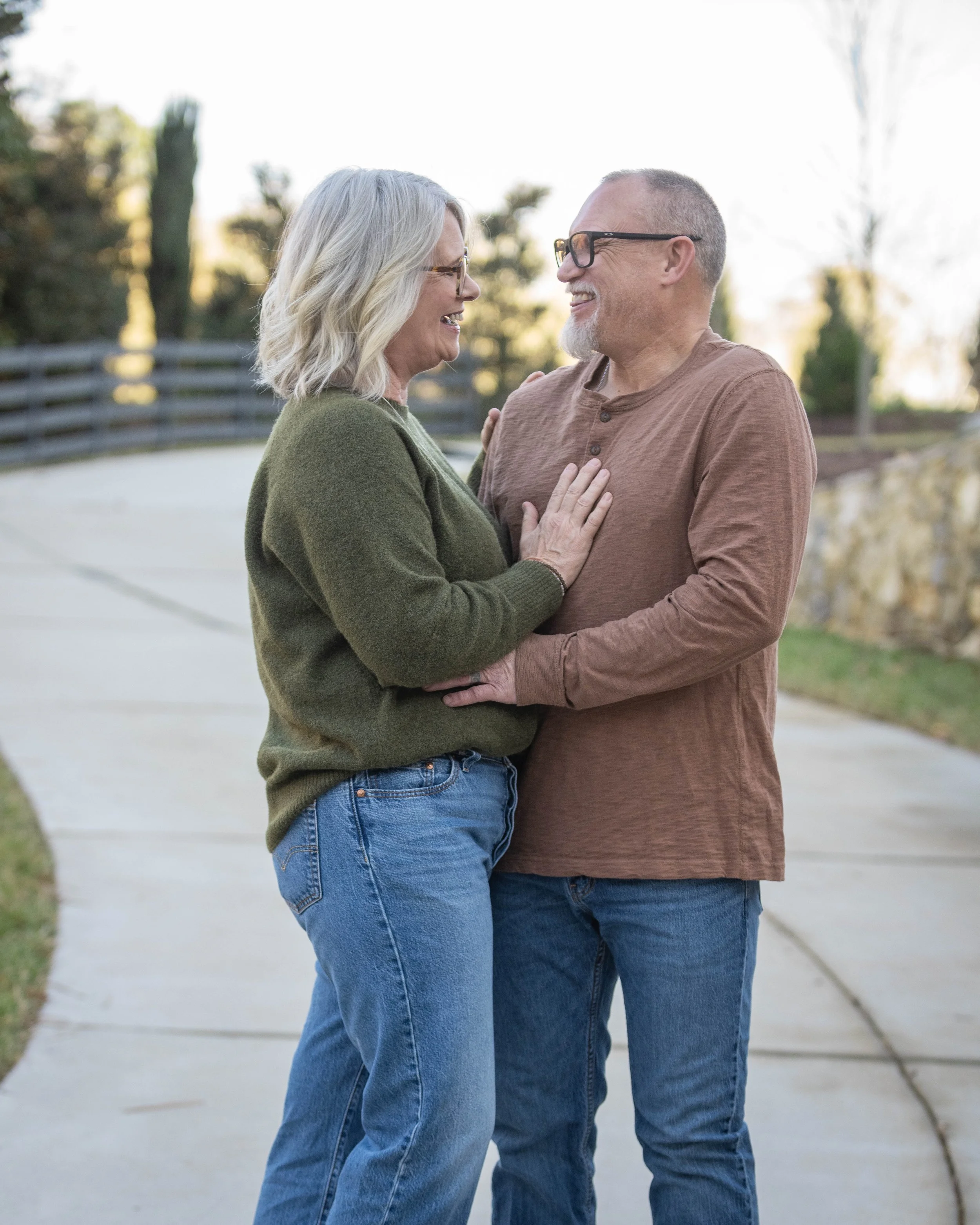 A middle-aged couple smiling and holding each other on a curved sidewalk outdoors during daytime.