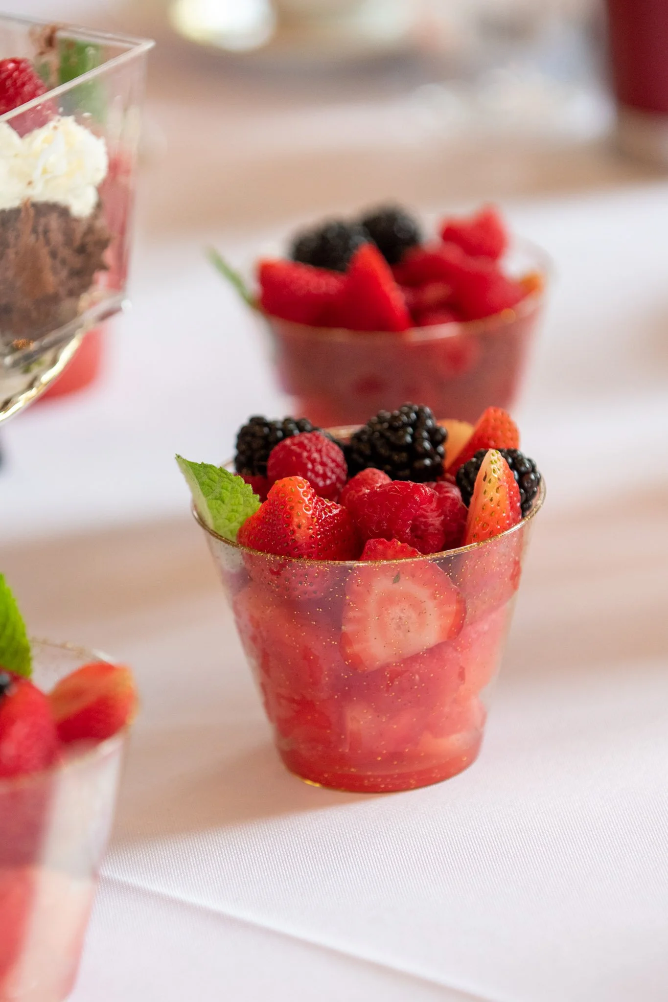 Clear cup filled with mixed strawberries, raspberries, blackberries, and mint leaves on a white tablecloth.