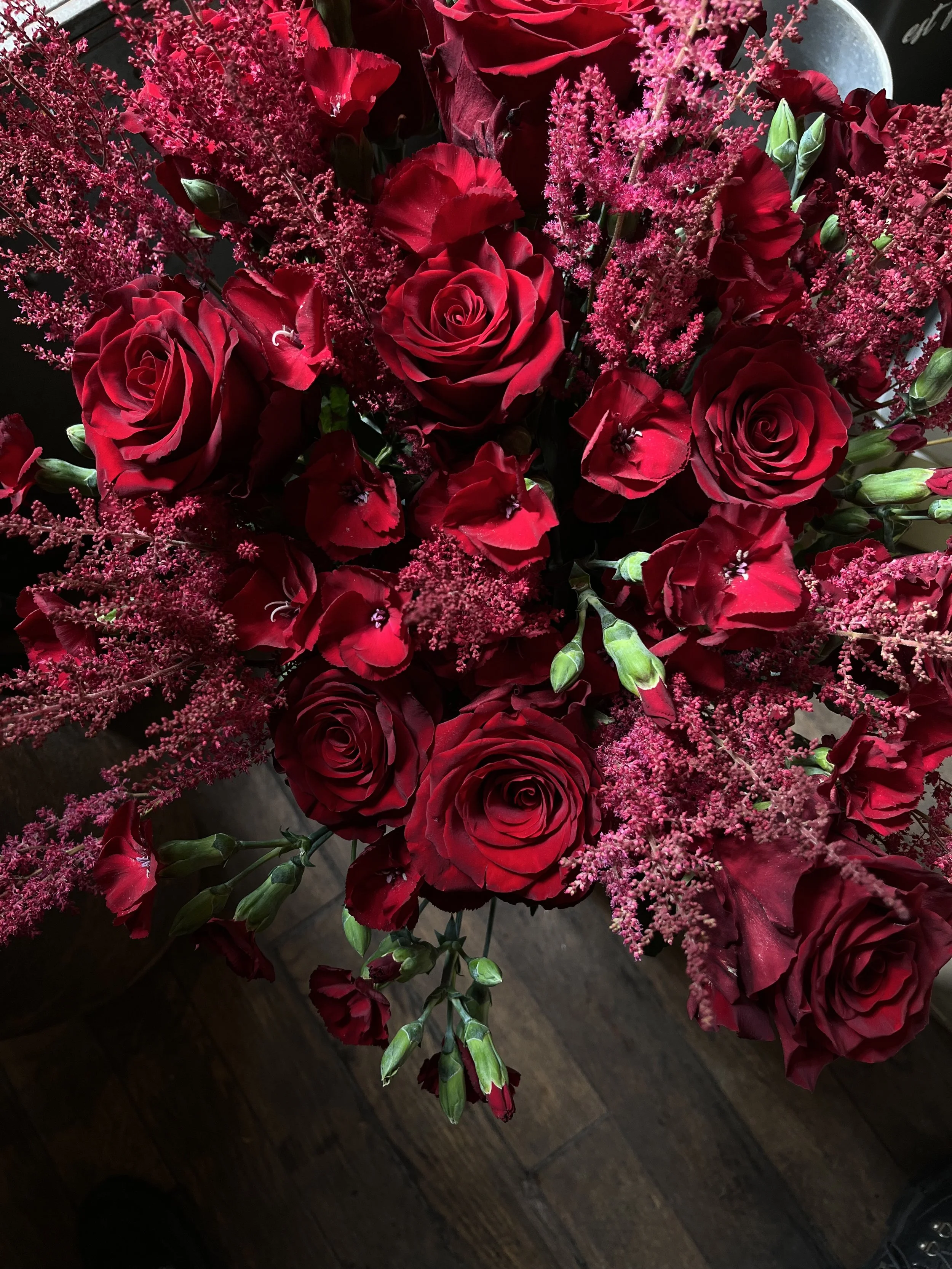 Un bouquet composé principalement de roses rouges, de fleurs de lys rose et de petites fleurs roses, sur un fond sombre.