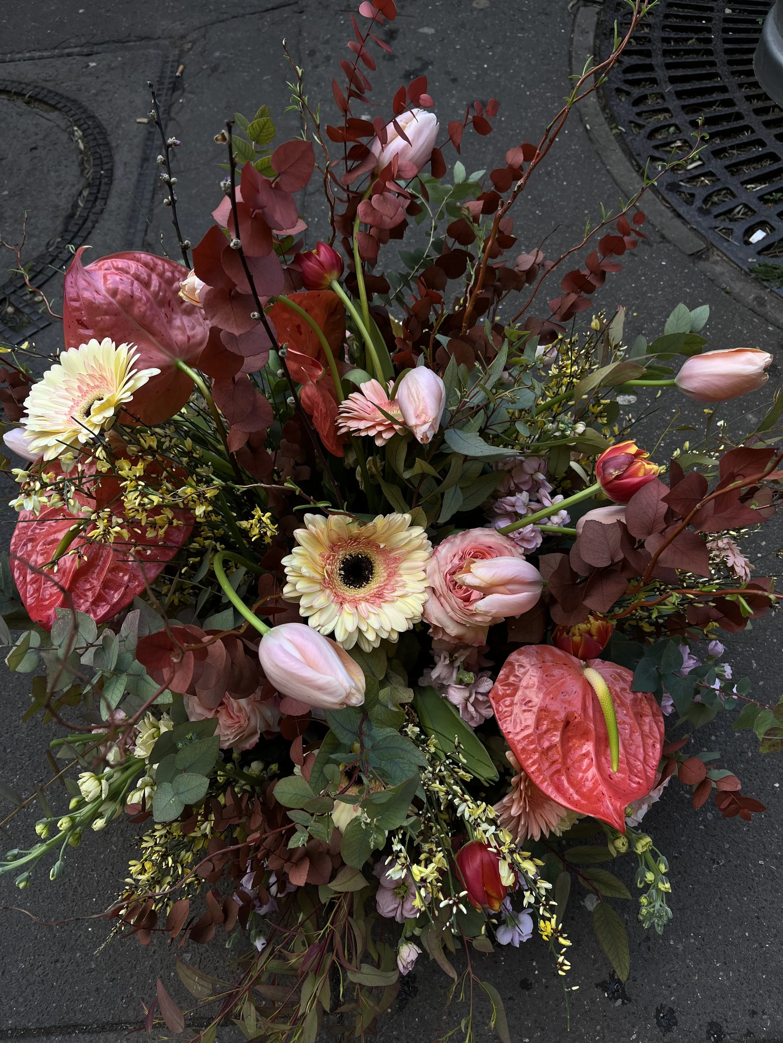 Un bouquet de fleurs colorées avec des roses, des tulipes, des gerberas, des anthuriums rouges, et différentes feuilles et branches.
