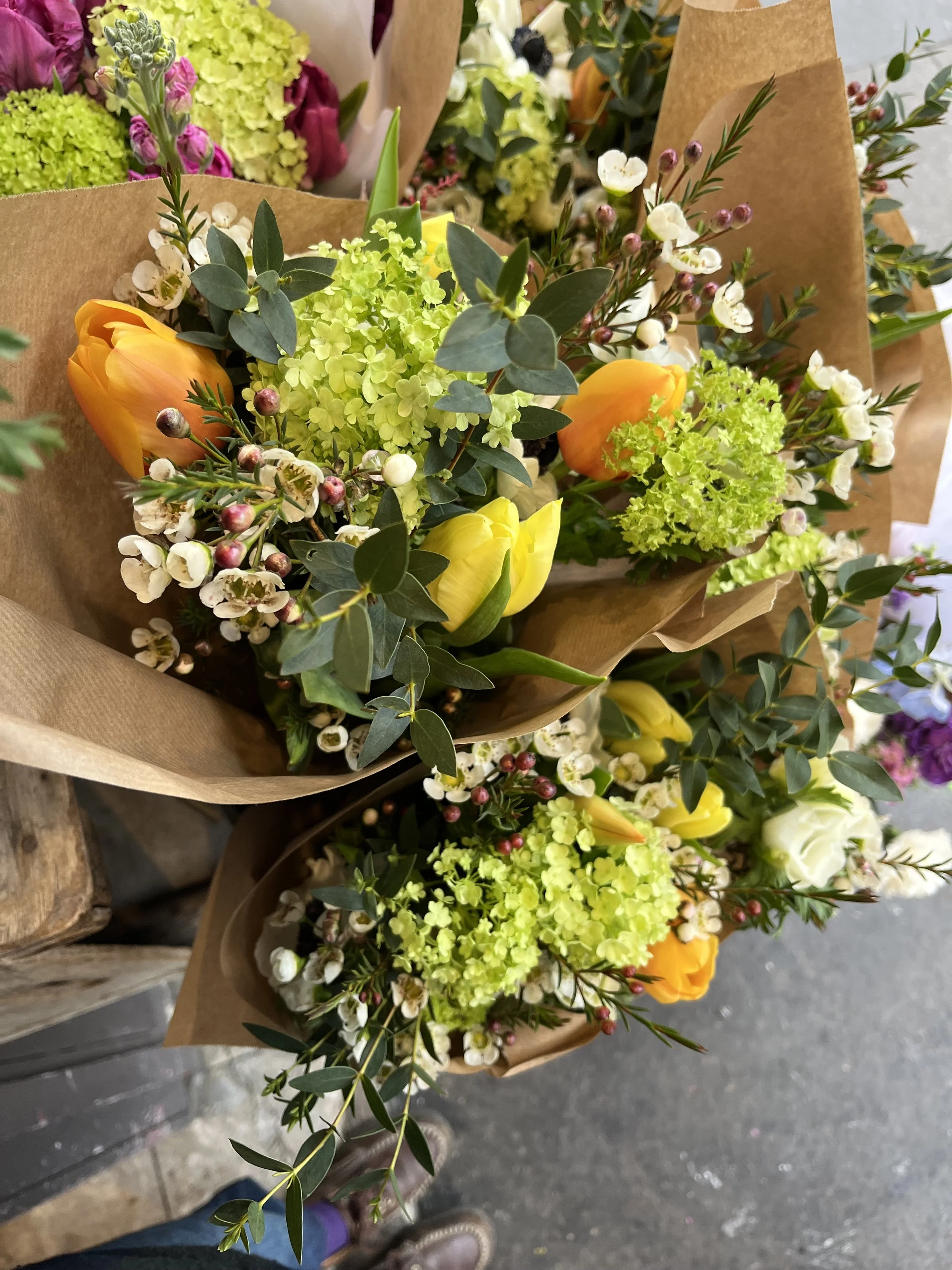 Bouquets de fleurs multicolores avec des tulipes jaunes et oranges, des hortensias vert clair, des houdes blanches, et diverses feuilles vertes, enroulées dans du papier craft.