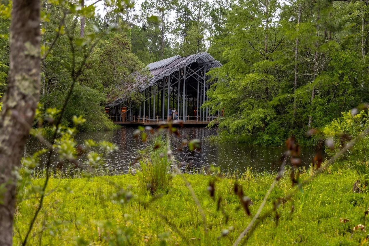 A covered wooden bridge over a pond in a lush green forest with trees and grass surrounding the water.